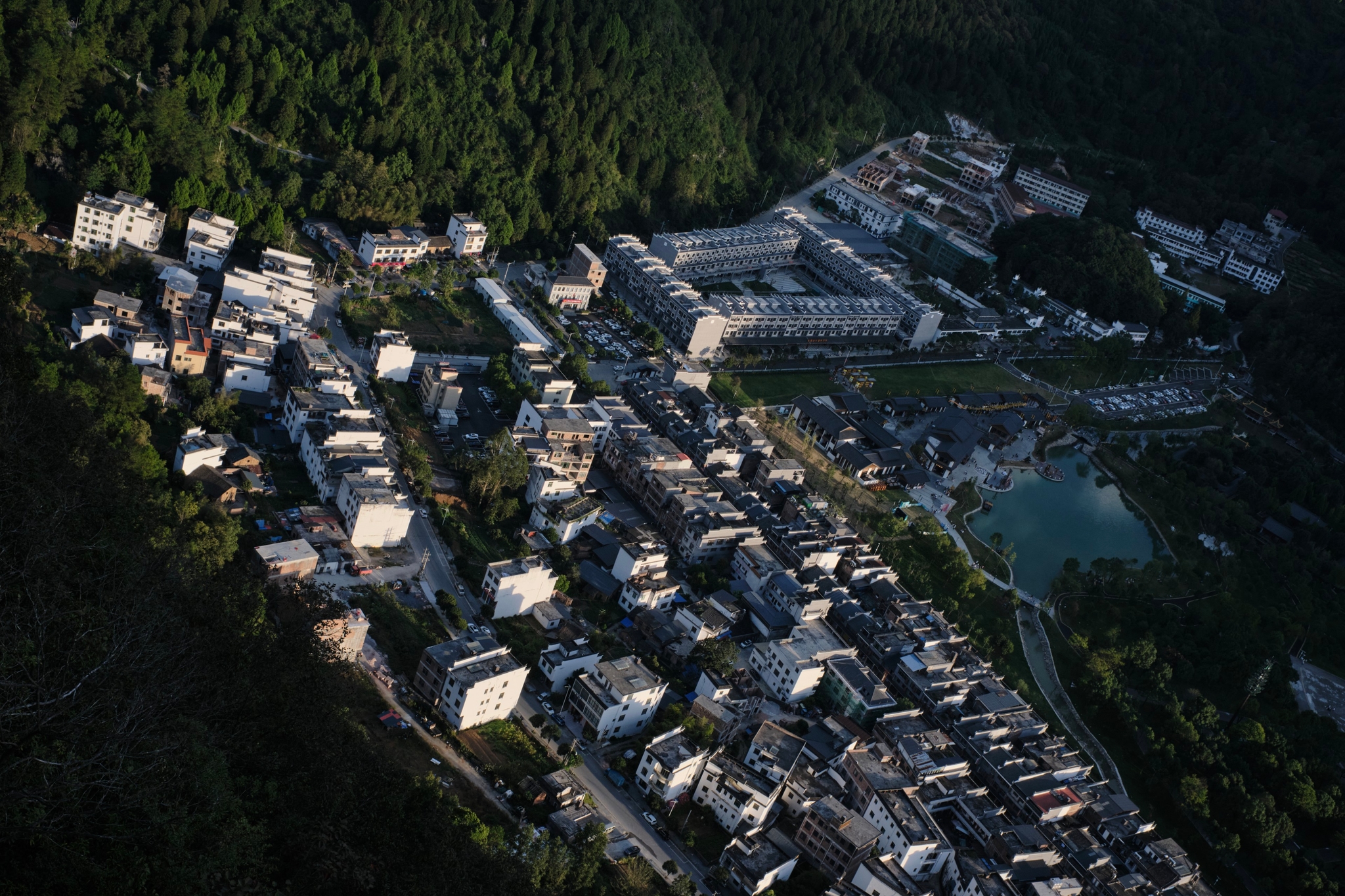 Aerial view of a village with numerous buildings bordered by a dark green forest, featuring a small body of water