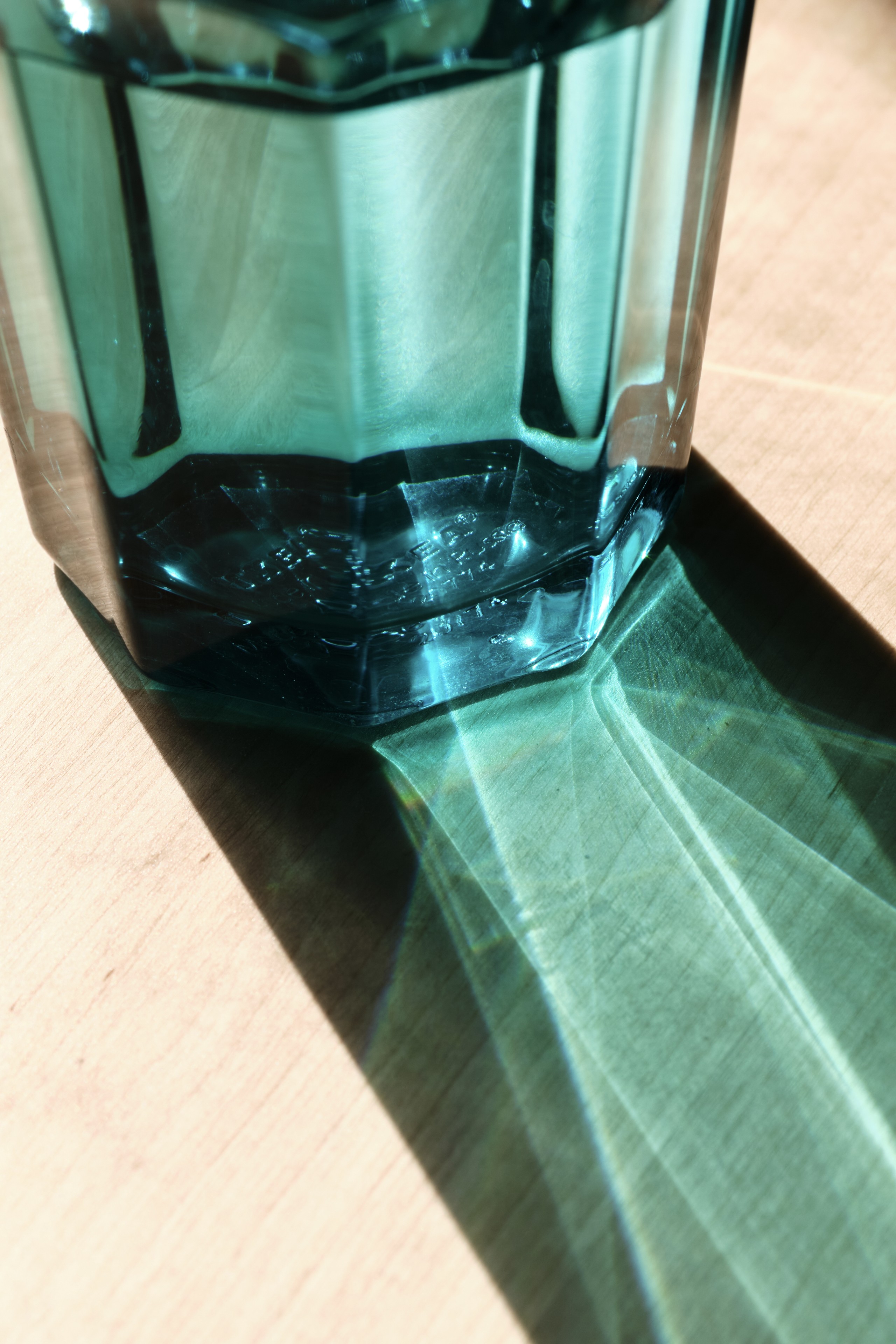 A faceted teal glass vessel sits on a light surface, casting a long, geometric shadow with distinct light refraction patterns