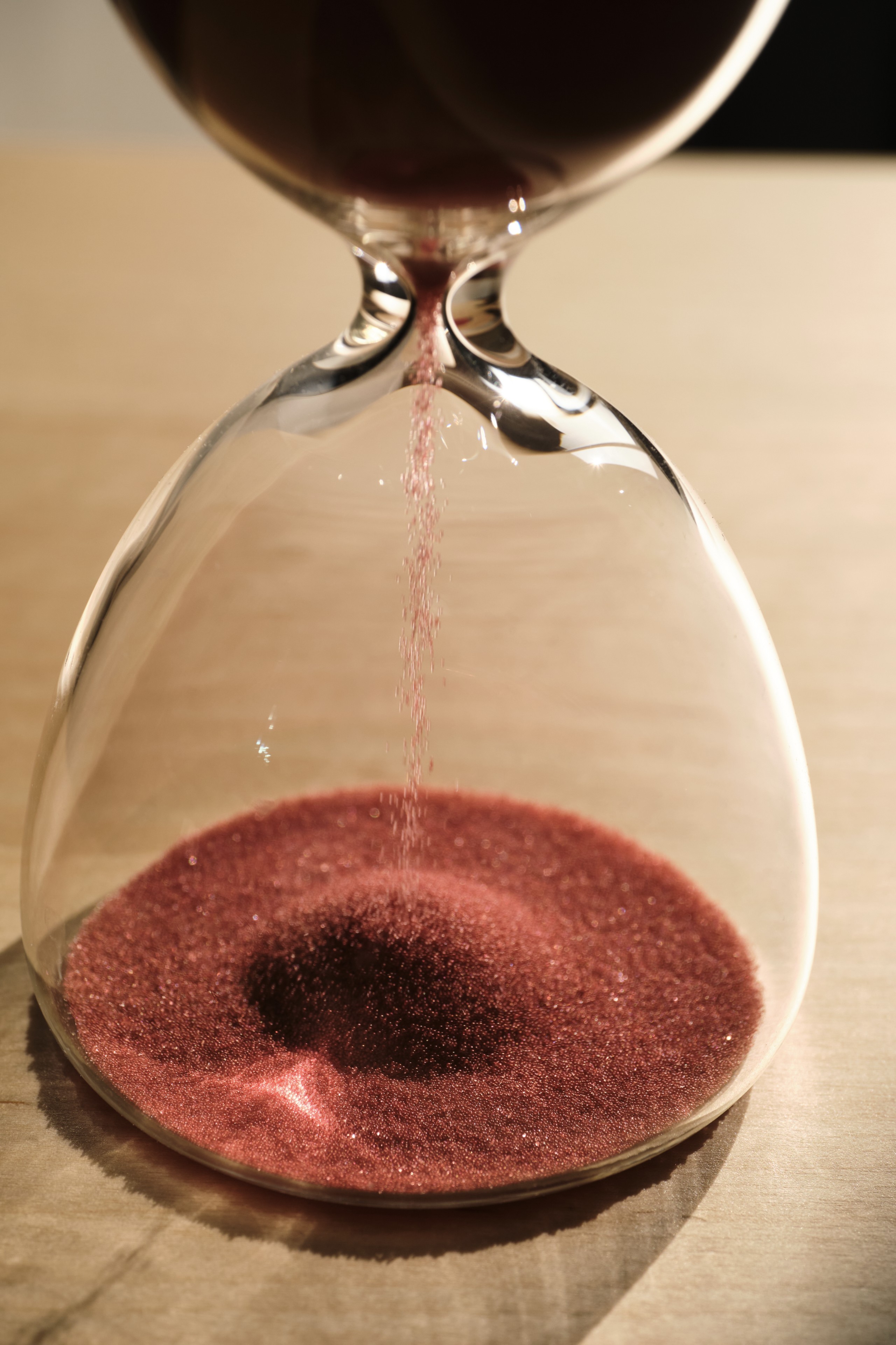 Close-up view of a glass hourglass with red sand grains falling through the narrow neck onto a mound forming in the lower bulb