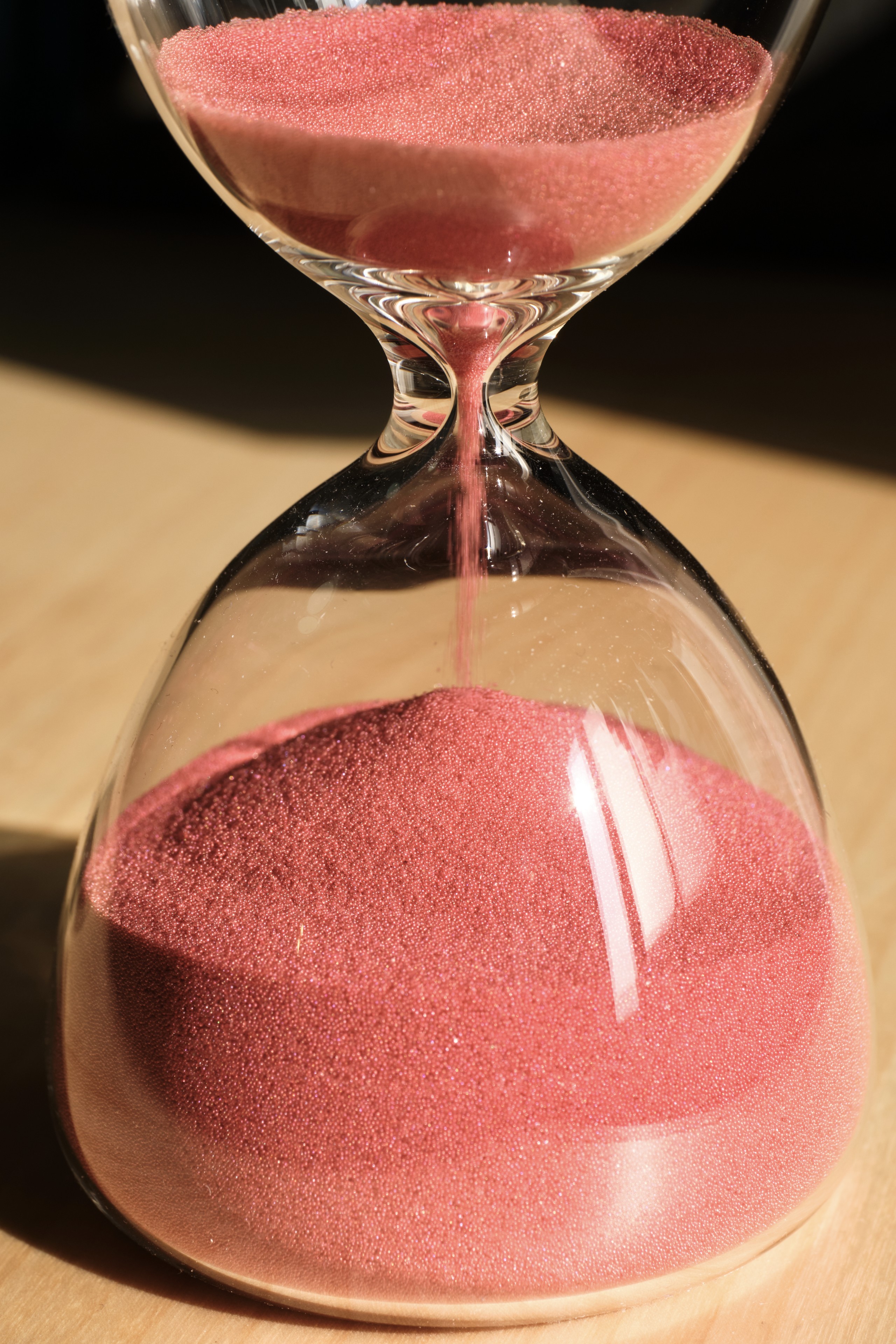 A glass hourglass filled with pink sand sits on a wooden surface, with sand flowing from the upper chamber to the lower chamber