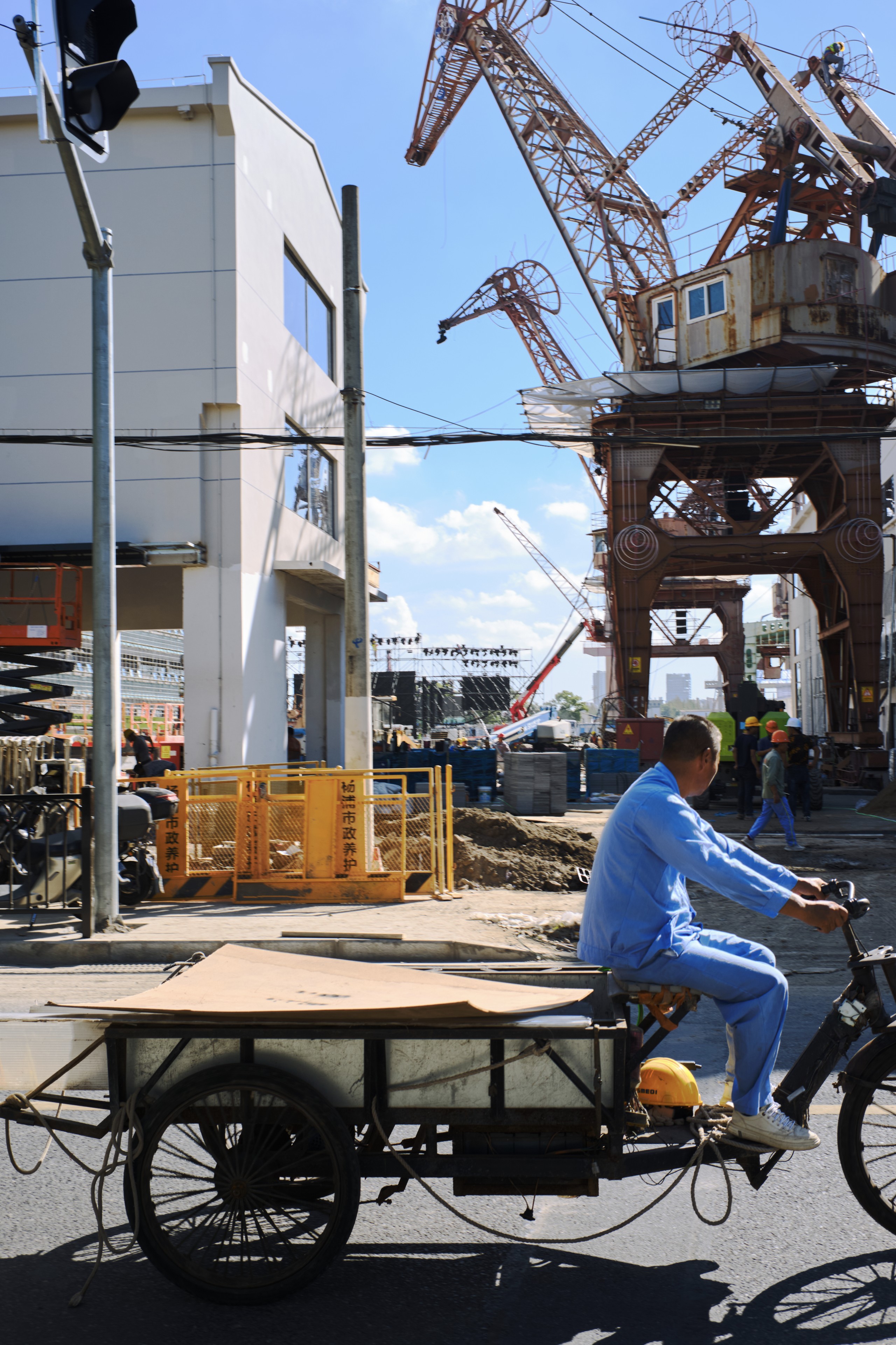 Man in blue uniform rides a cargo bicycle past large rusted industrial cranes, a white building, and a construction site under a clear blue sky