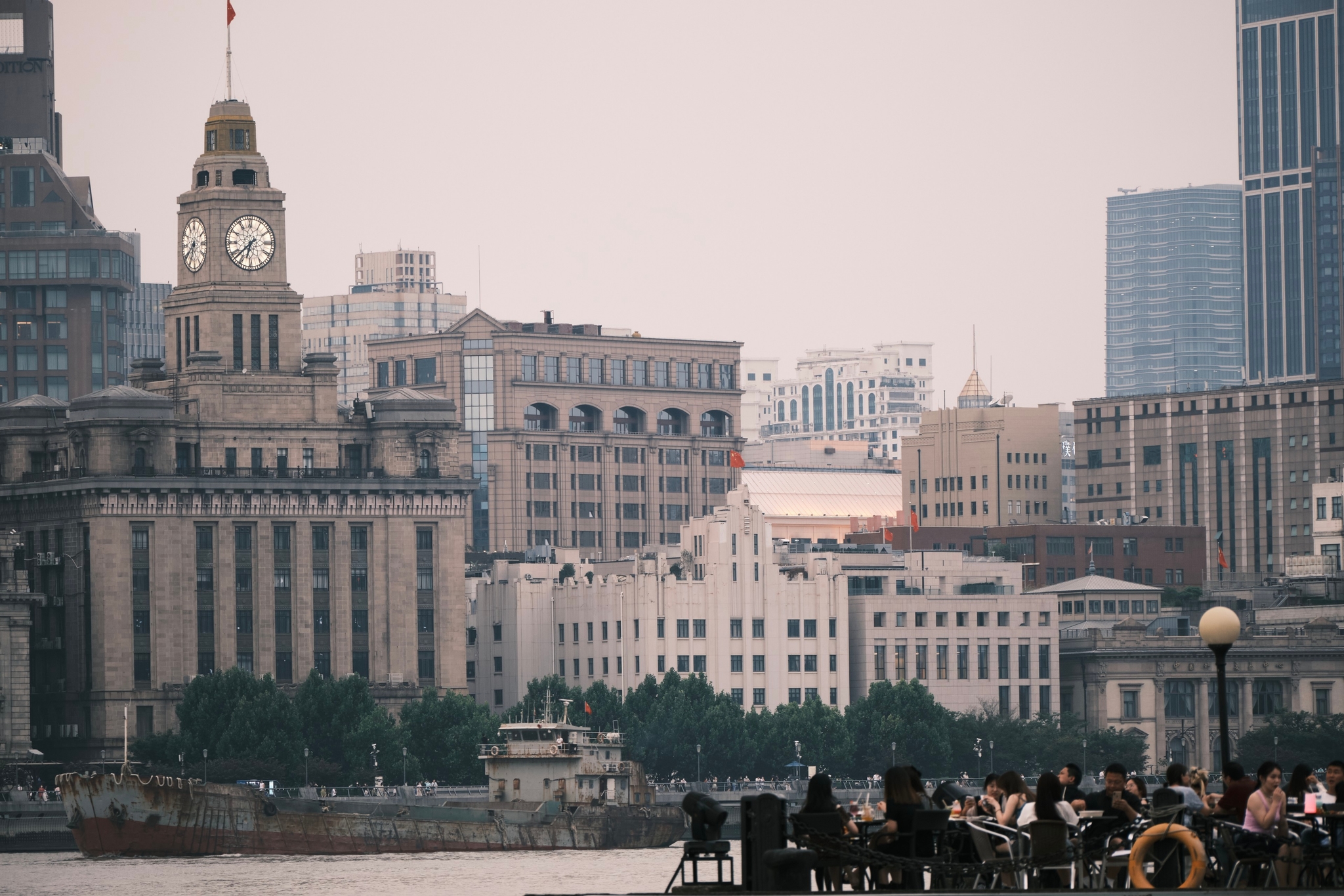 A riverside view of the Shanghai Bund featuring colonial-era architecture with a prominent clock tower and modern skyscrapers in the background, with figures silhouetted along the waterfront promenade