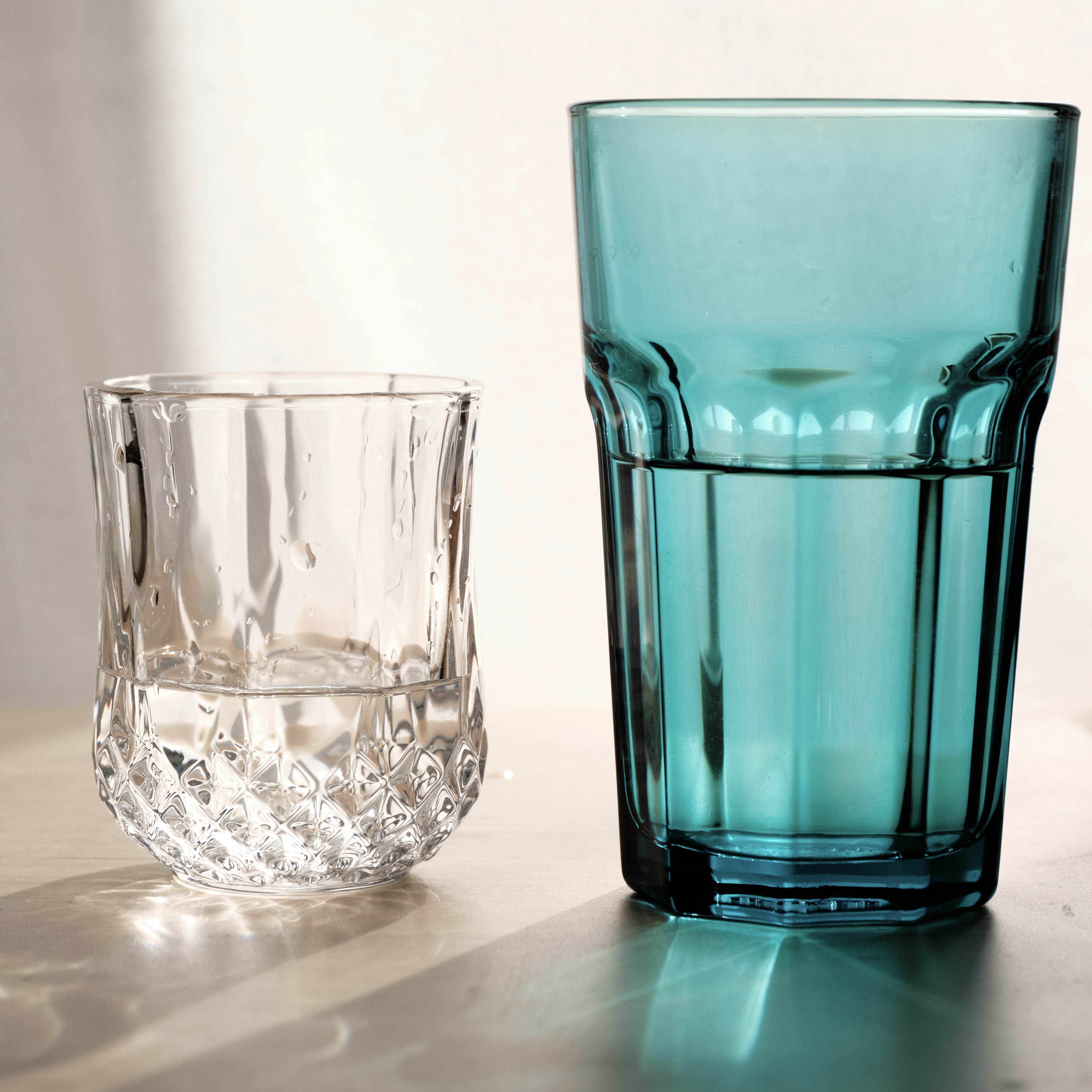 A small textured clear glass sits next to a larger teal fluted glass, both partially filled with water, illuminated by soft sunlight casting reflections on a neutral surface