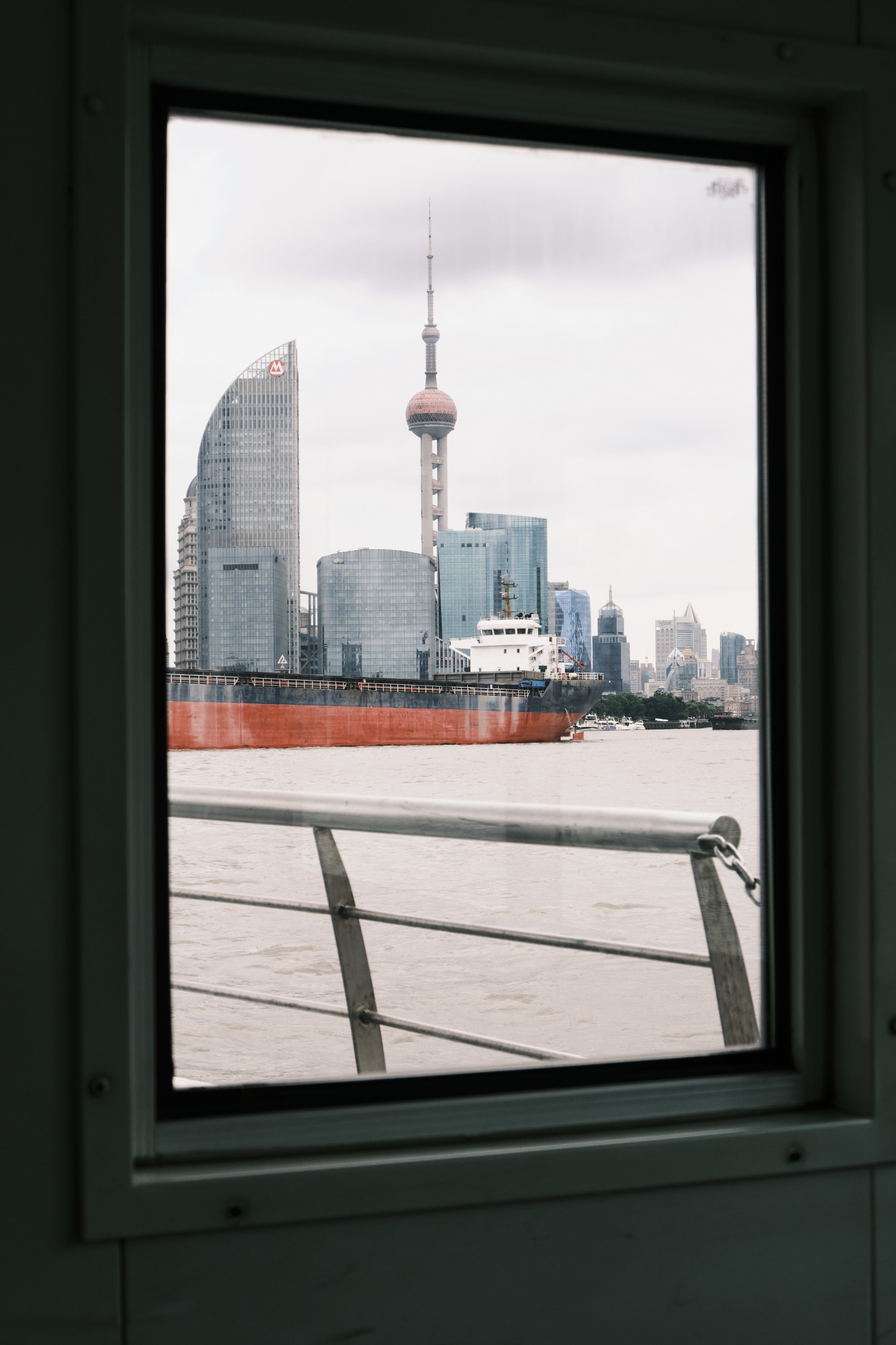 A rectangular window frame looking out over a river towards the Shanghai skyline, featuring the Oriental Pearl Tower and a large orange cargo ship passing in front of skyscrapers under a cloudy sky