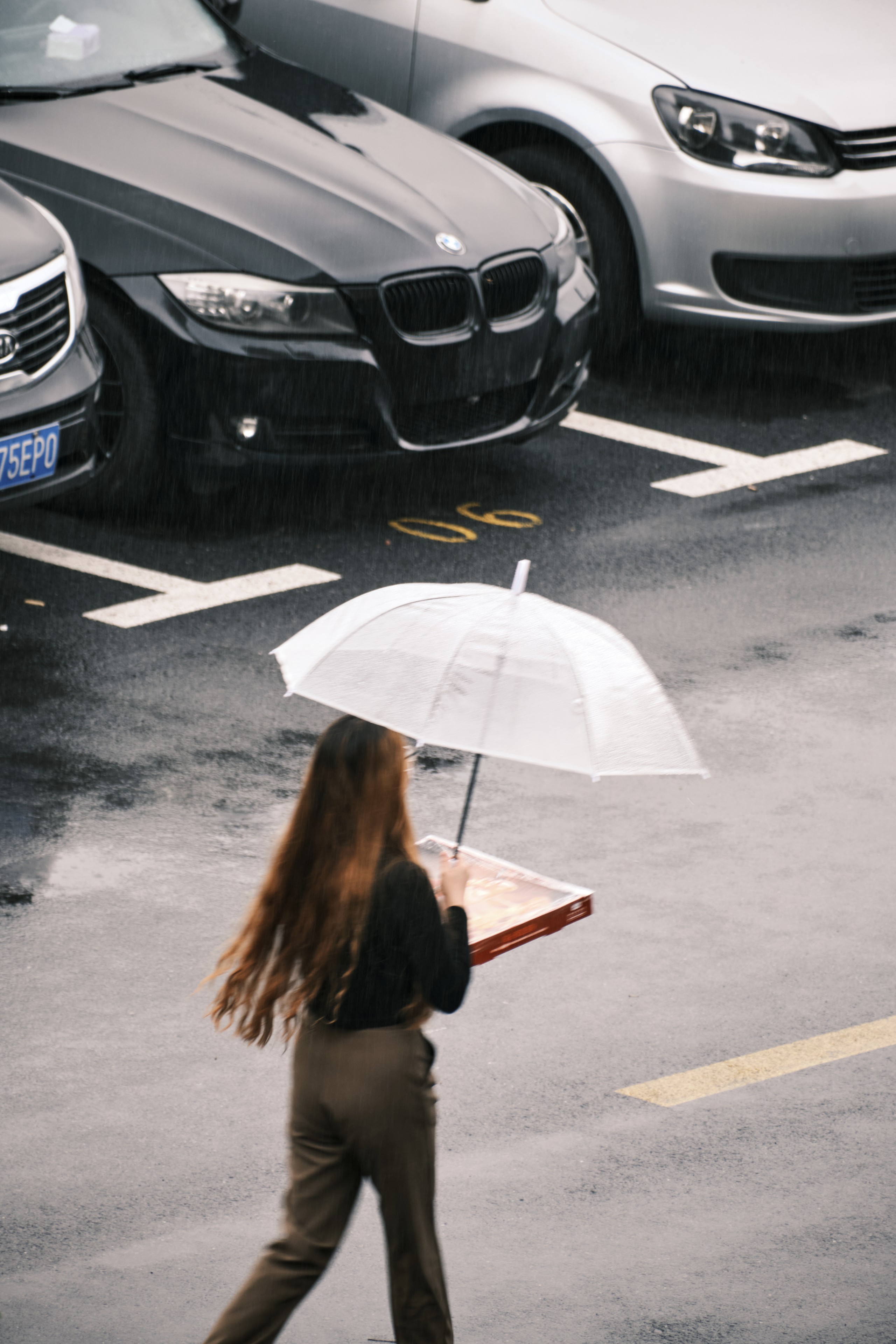 A woman with long brown hair carries a pizza box and a clear umbrella while walking across a wet, marked parking lot in front of two parked cars