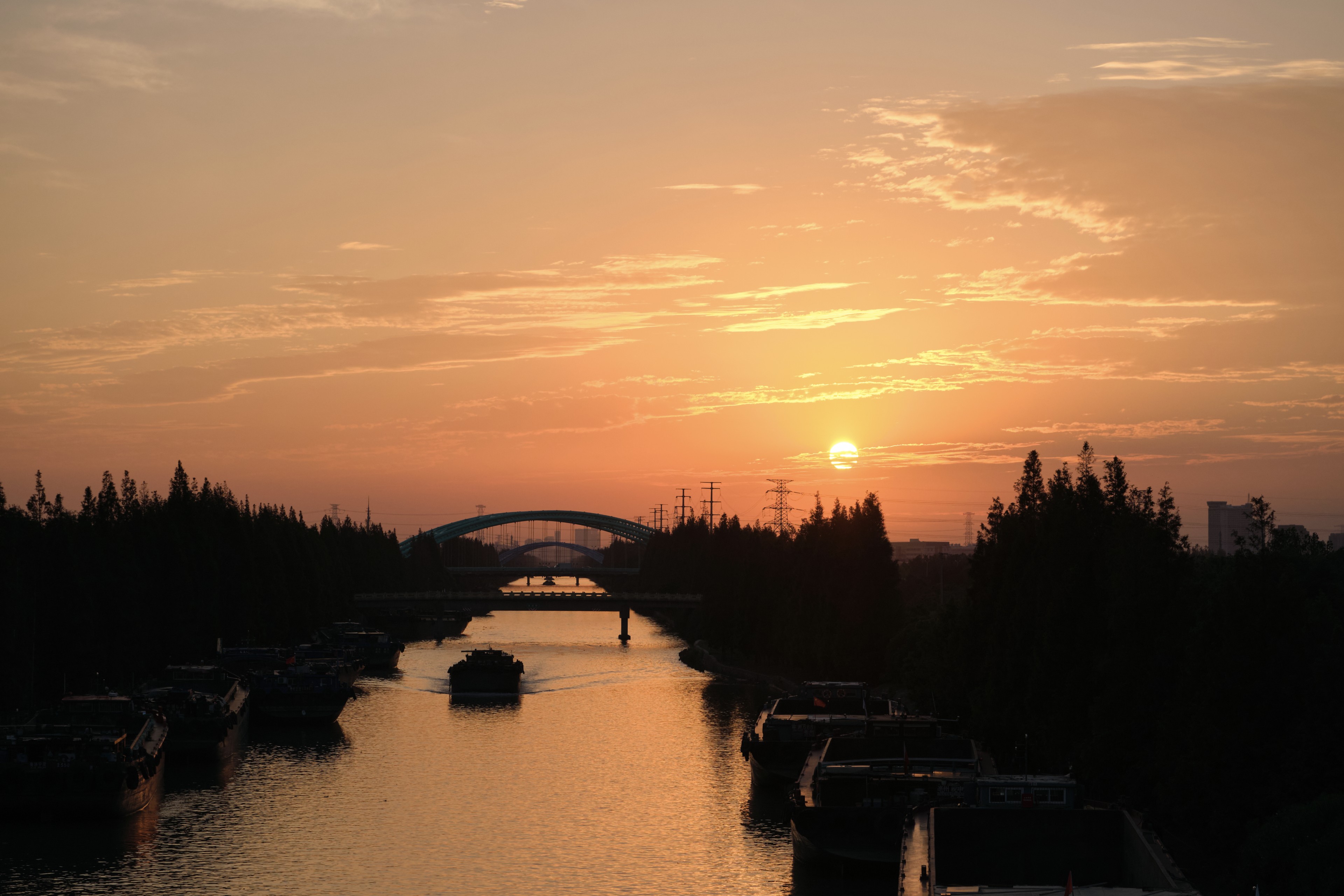A wide angle sunset view over a river with several silhouettes of moored and traveling boats, framed by dark tree lines and distant bridge arches