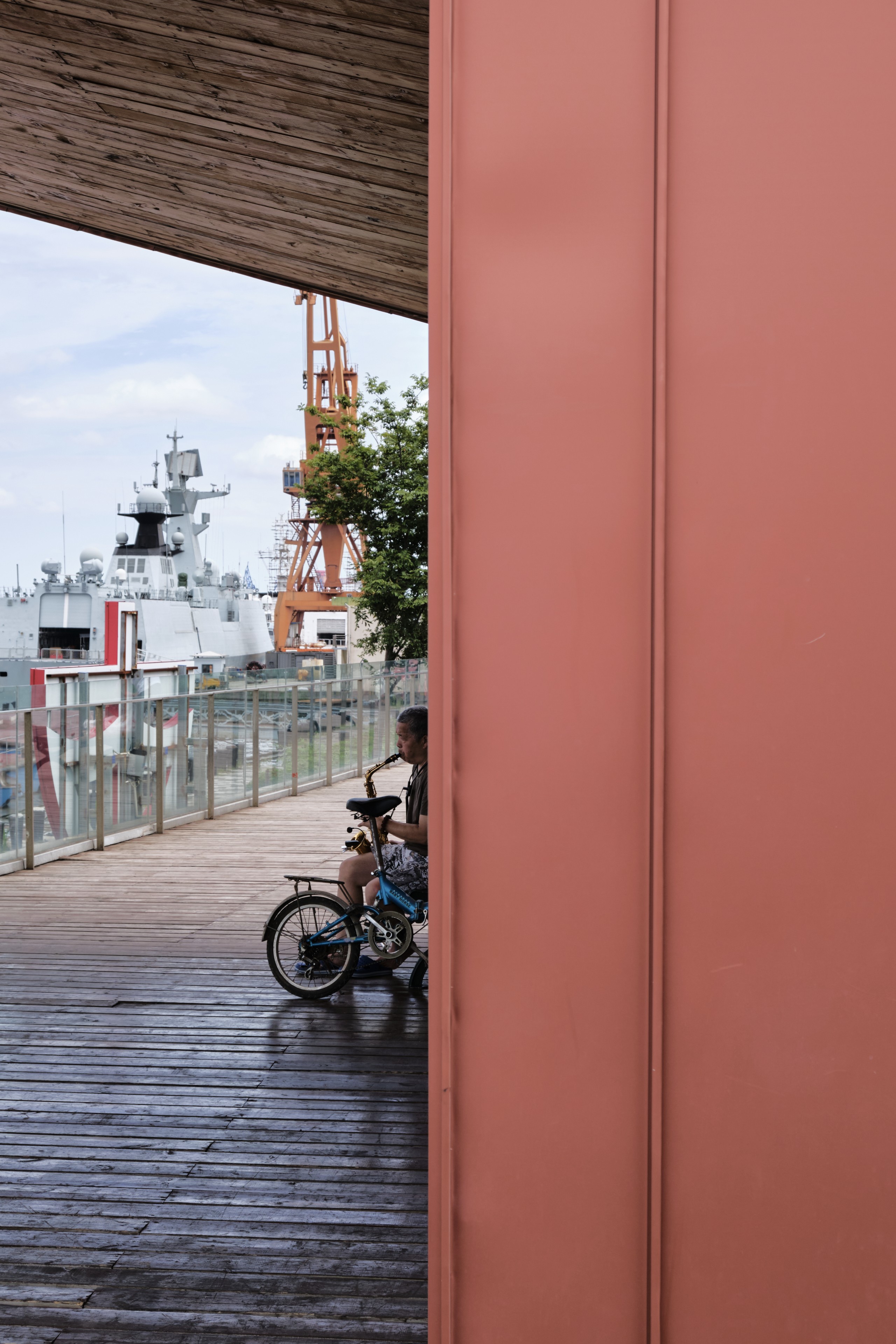 A person on a bicycle sits on a wooden pier near a railing with a large ship and orange industrial crane in the background, partially obscured by a red wall in the foreground