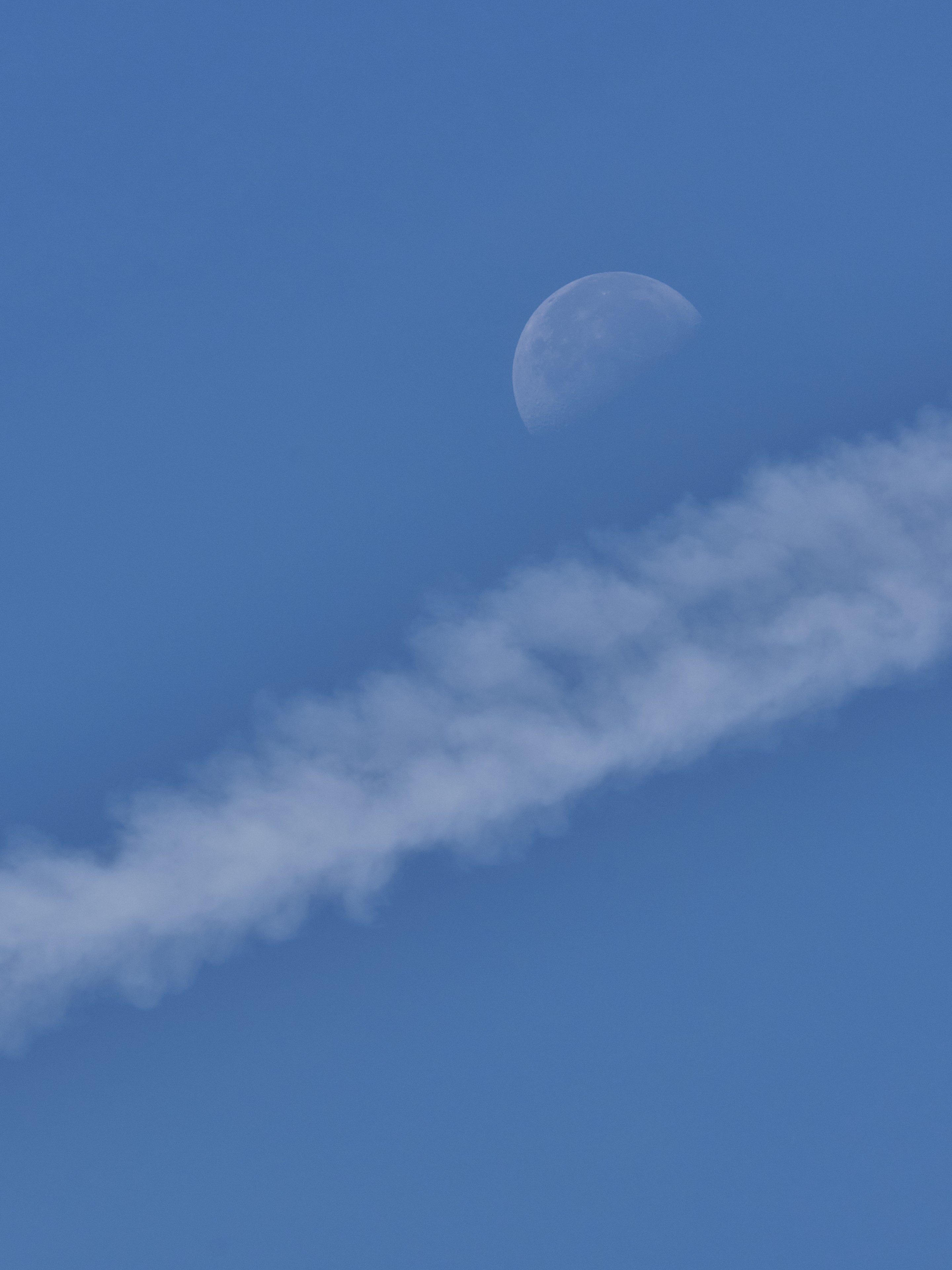 A waxing gibbous moon positioned in a clear blue sky above a diagonal contrail of white cloud vapor
