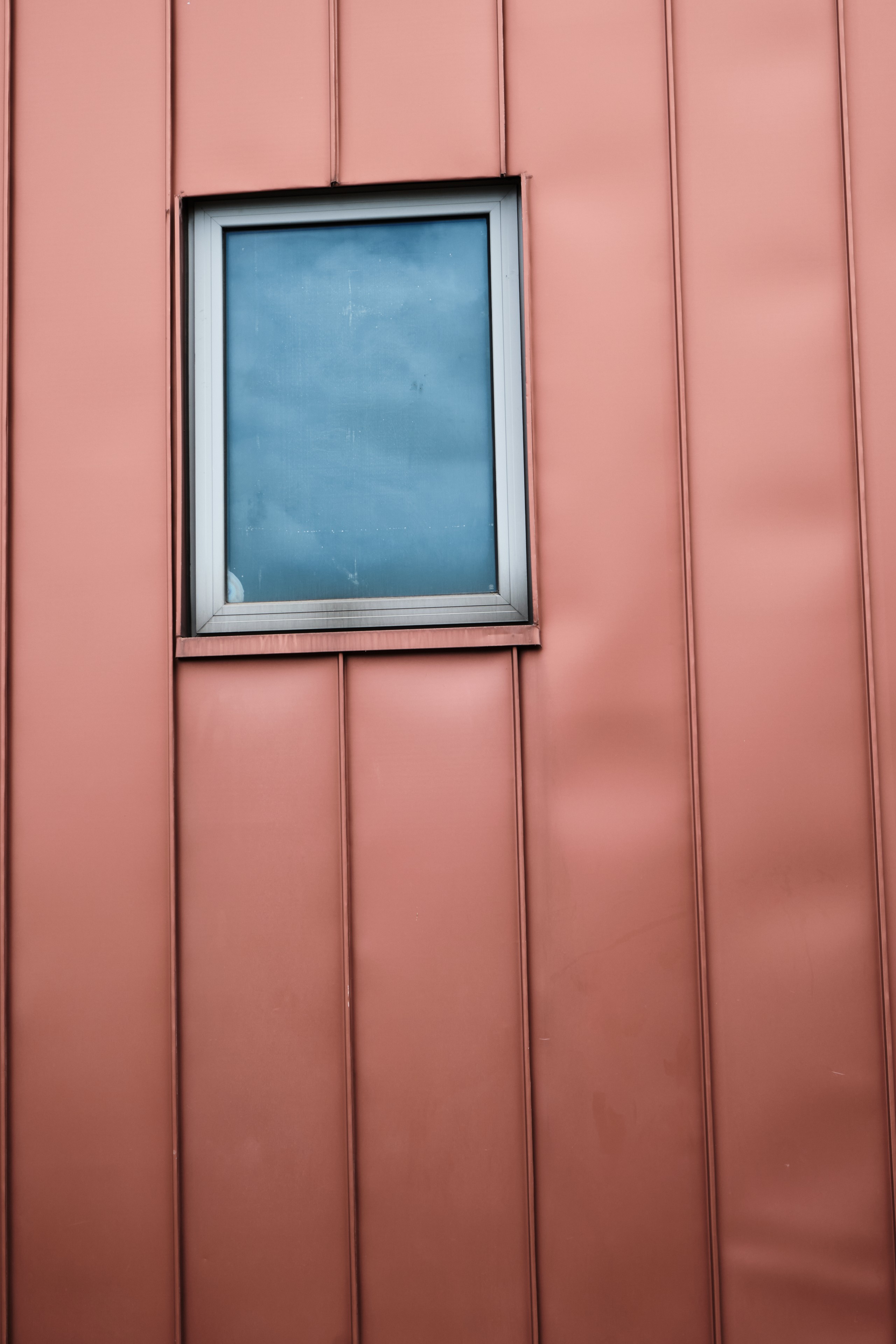A rectangular window set into a vertical panelled wall with a dusty rose metallic finish, reflecting a cloudy blue sky
