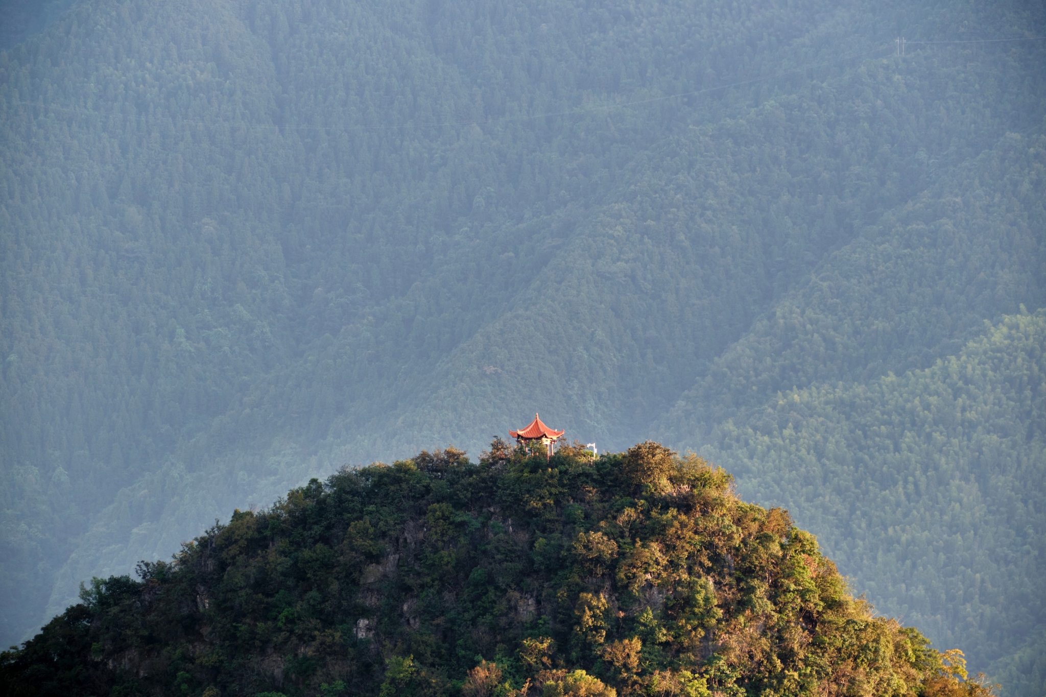 A small orange-roofed structure sits atop a densely forested mountain peak against a backdrop of hazy, layered blue mountain ridges