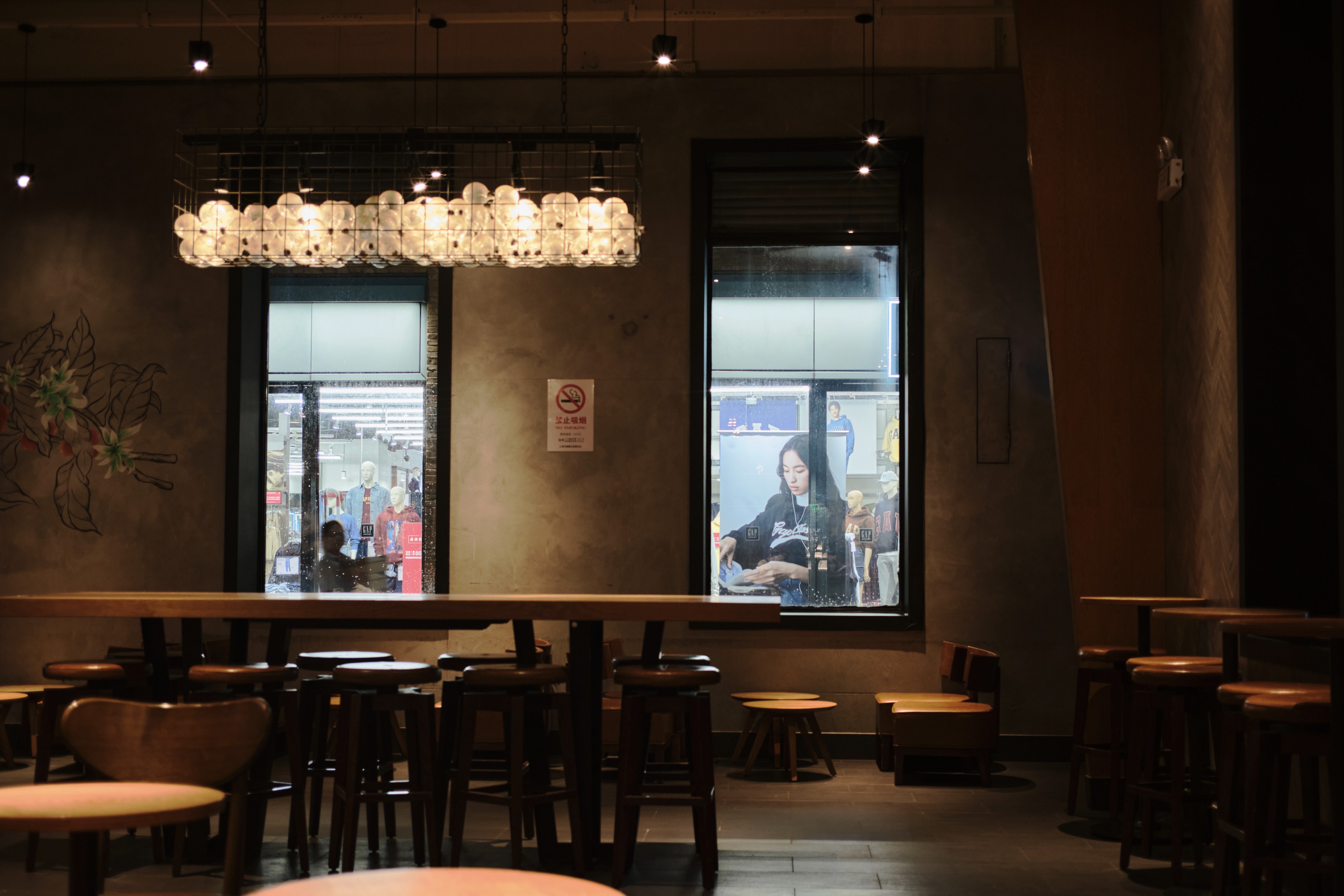 Dimly lit cafe interior with a long communal table, two windows revealing an urban scene, and a decorative chandelier