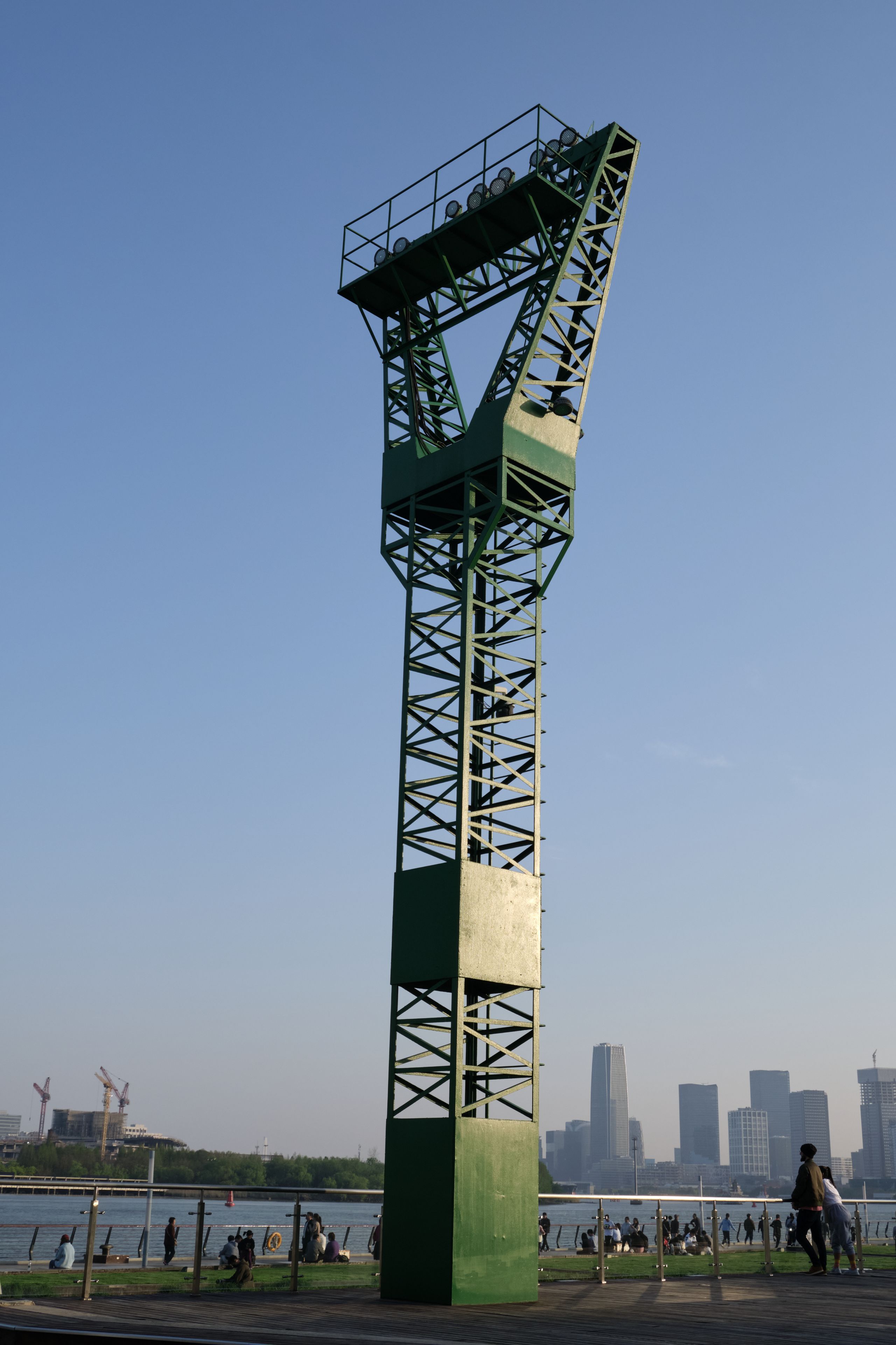 Green metal lattice tower, clear blue sky, waterfront with distant cityscape and people