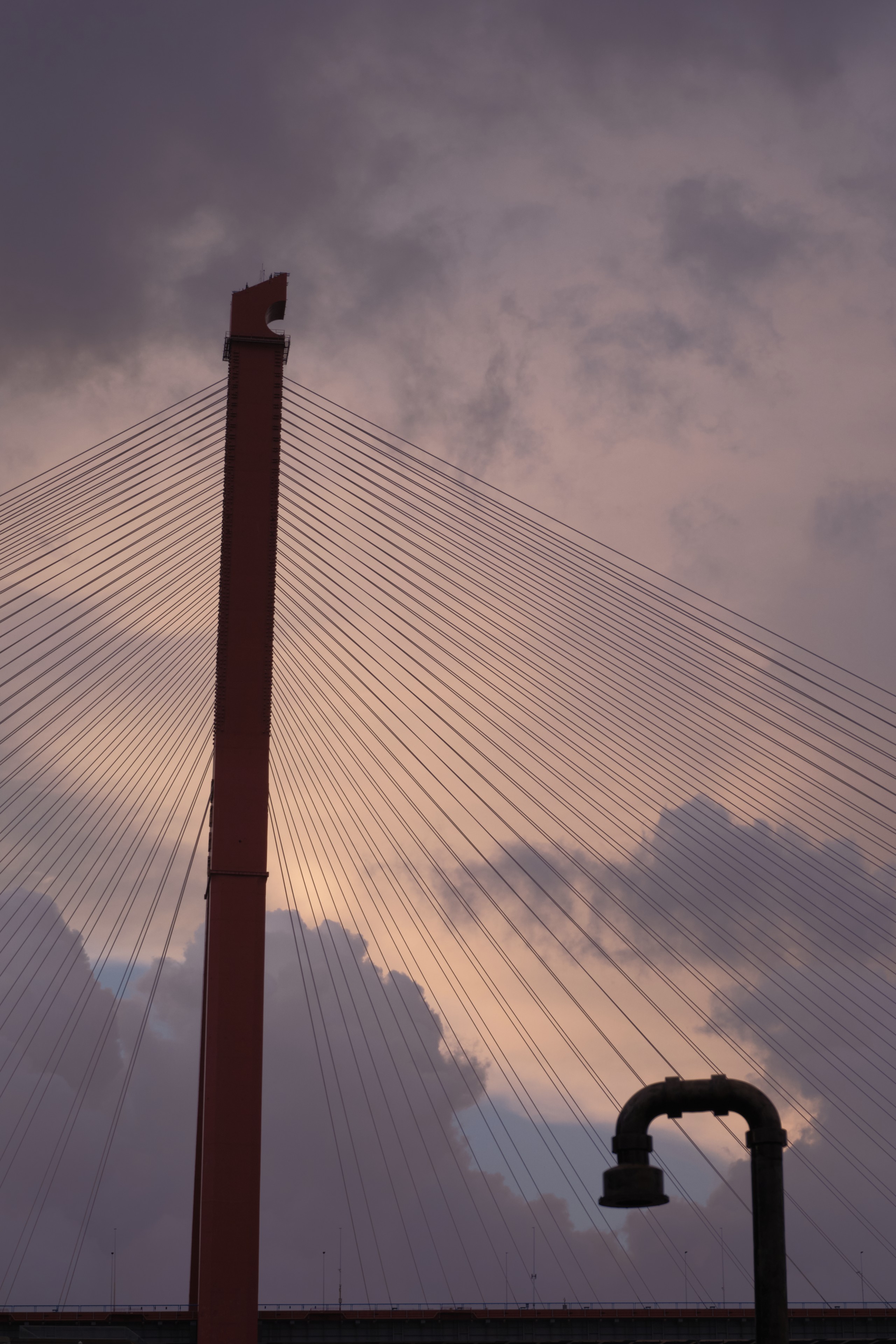 Red cable-stayed bridge pylon with radiating cables against a cloudy, multi-hued sky. Dark L-shaped pipe structure in foreground