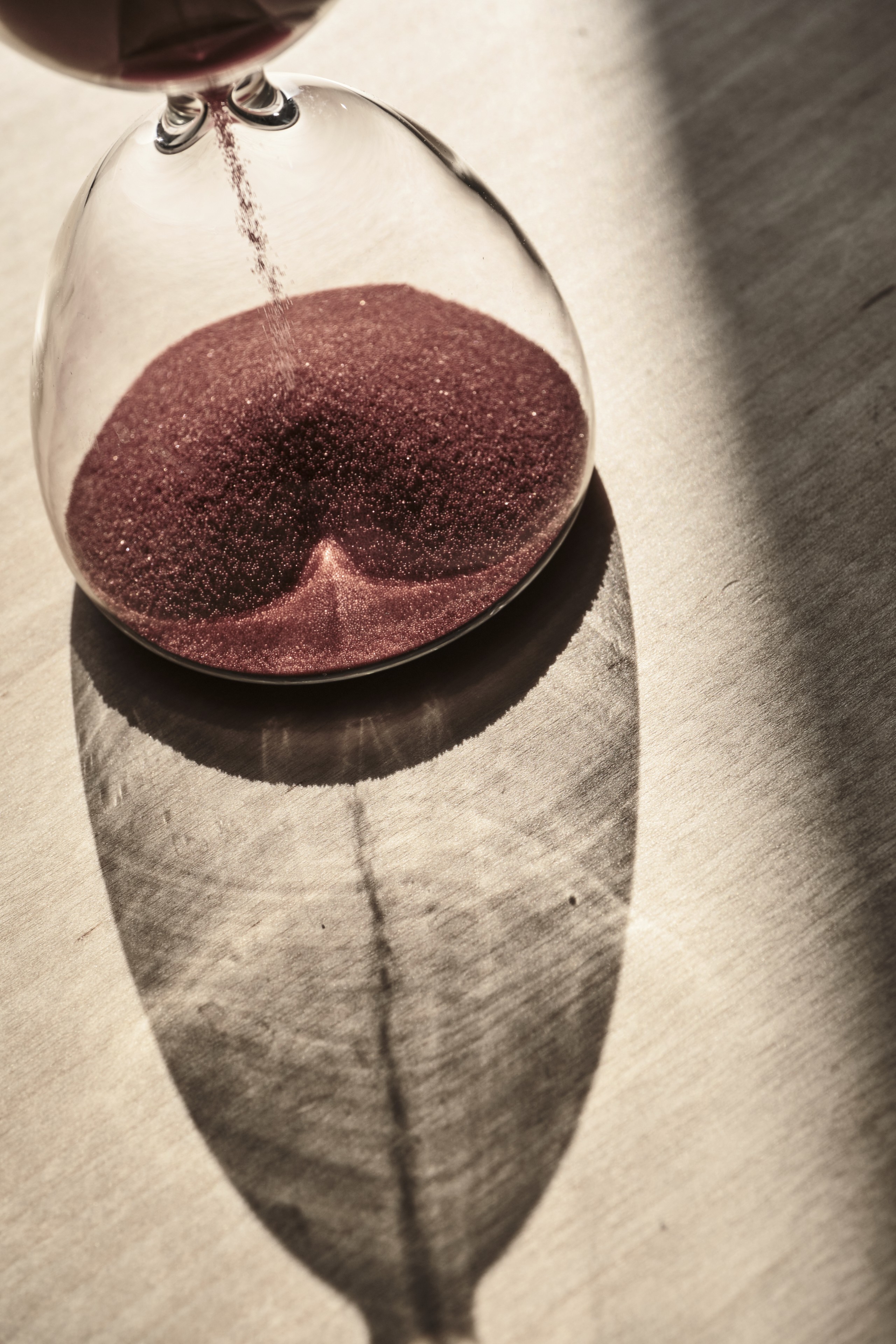 A close-up view of a glass hourglass filled with red sand sitting on a textured surface, casting a long, dark shadow in bright natural light