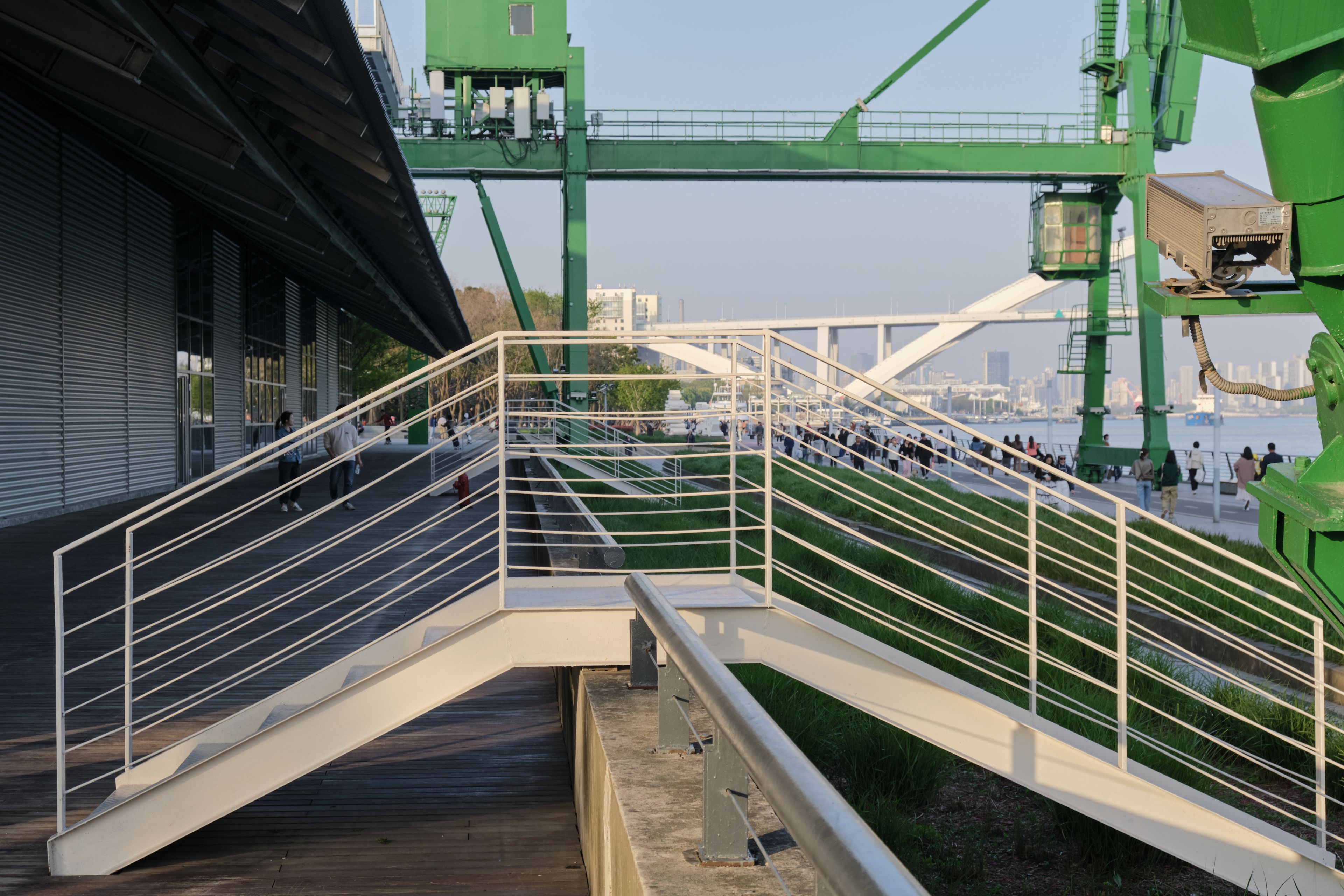 Modern metal staircase leading to waterfront, green industrial cranes, white arched bridge, building on left