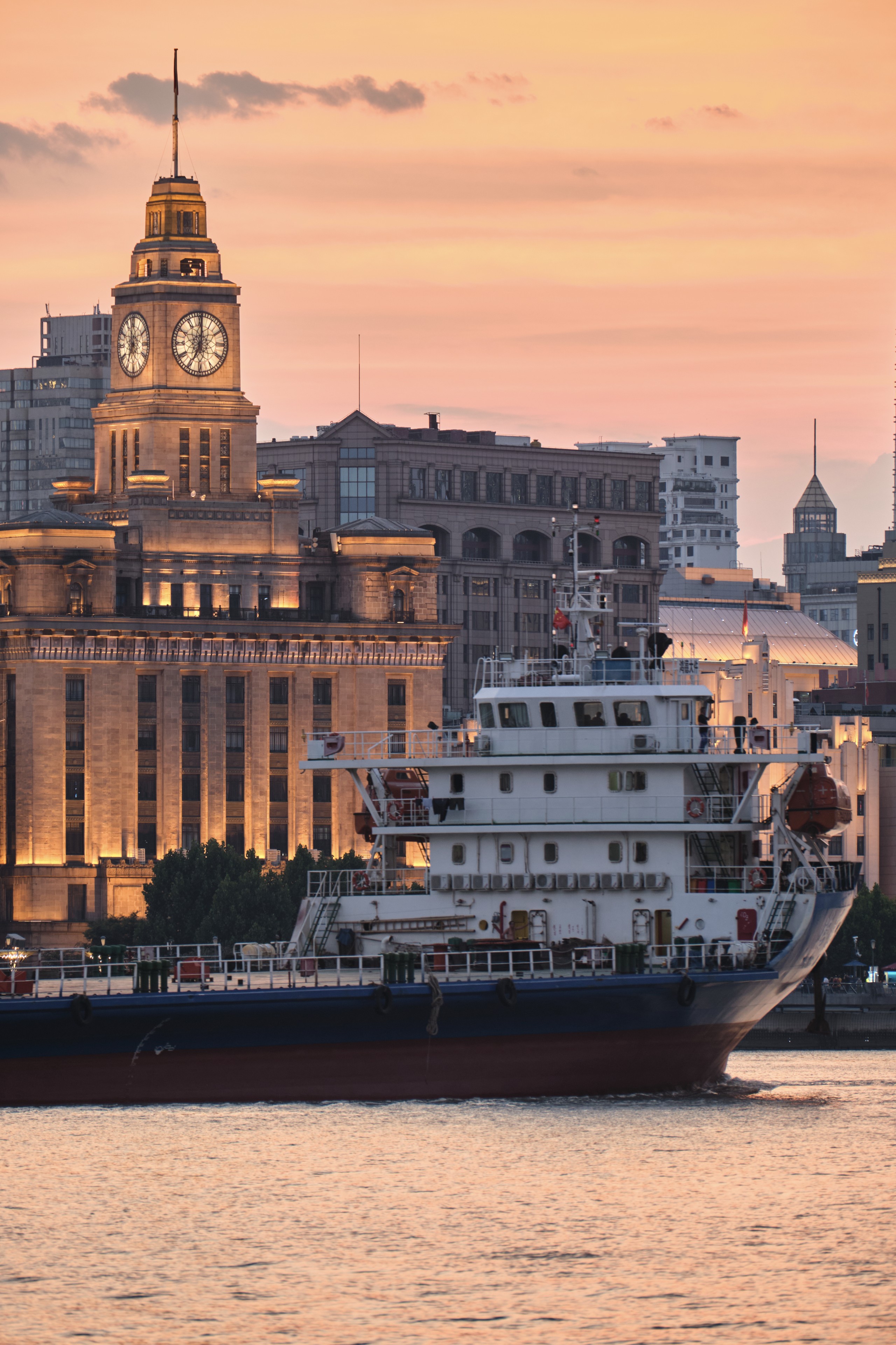 A cargo ship cruises along the Huangpu River in Shanghai past the historic clock tower of the Customs House during sunset with soft orange light reflecting on the water