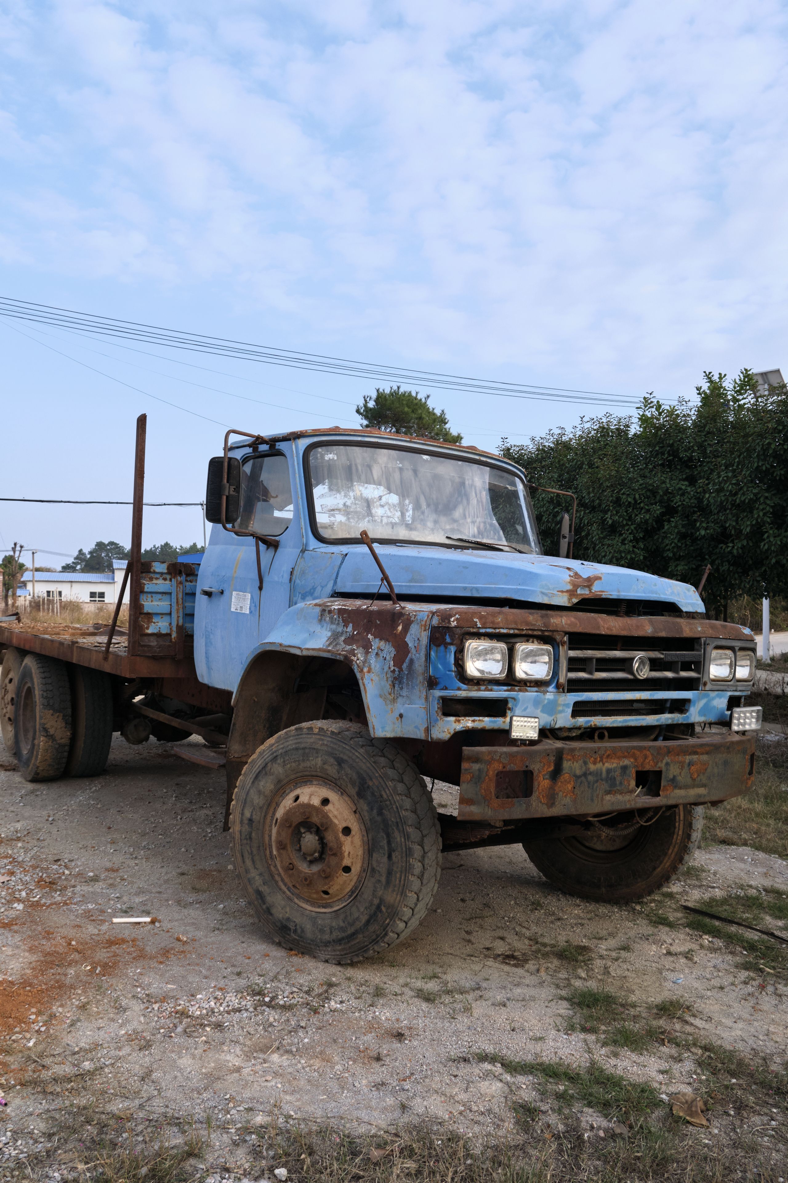 A faded blue, rusty, vintage flatbed truck is parked on a dirt lot under a cloudy sky, with hints of green foliage and structures in the background