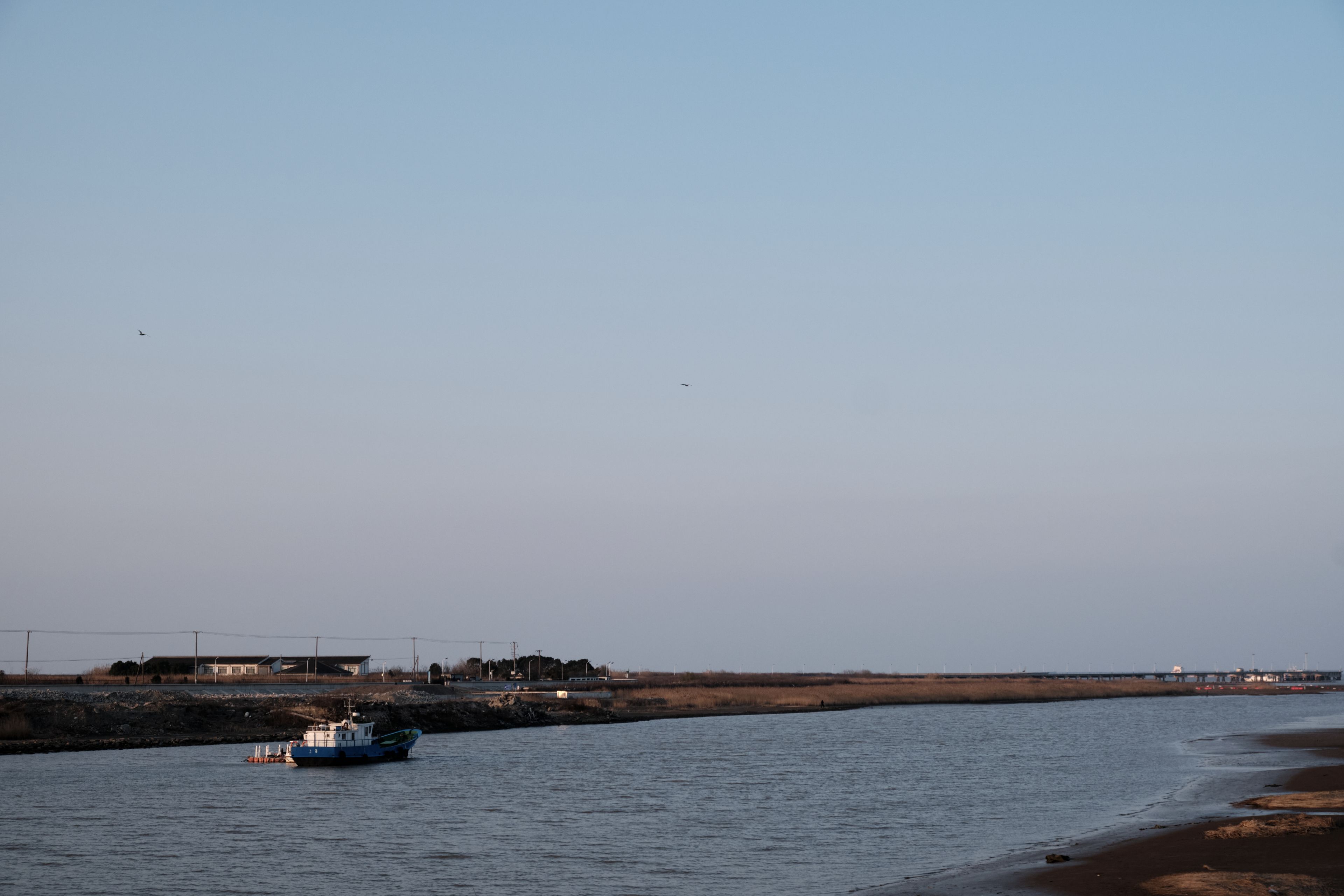 A small blue and white boat floats on a calm river. Distant land with buildings and vegetation lines the left bank under a clear blue sky