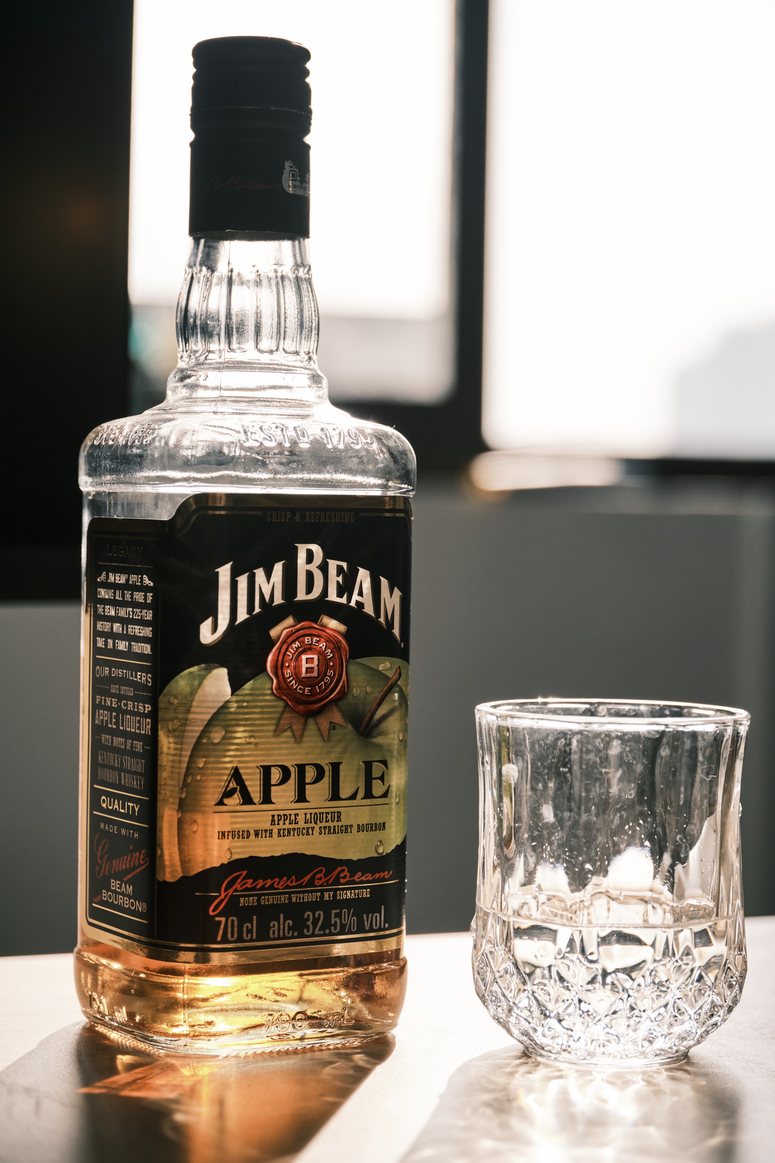 A bottle of Jim Beam Apple whiskey stands next to an empty crystal glass on a reflective surface with a window in the background
