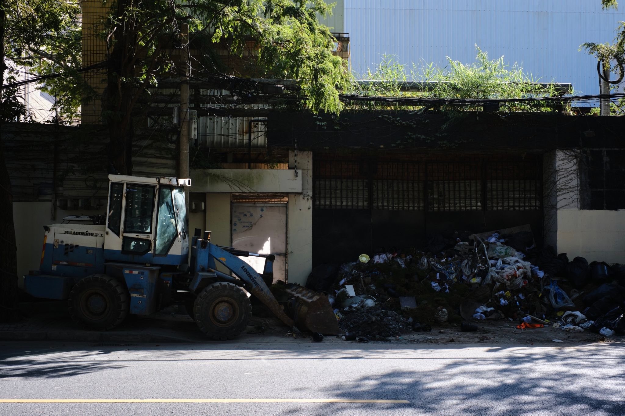 A blue and white front-end loader is positioned next to a large pile of dark, lumpy material on an asphalt road, with a dark wall and green foliage in the background under sunlight