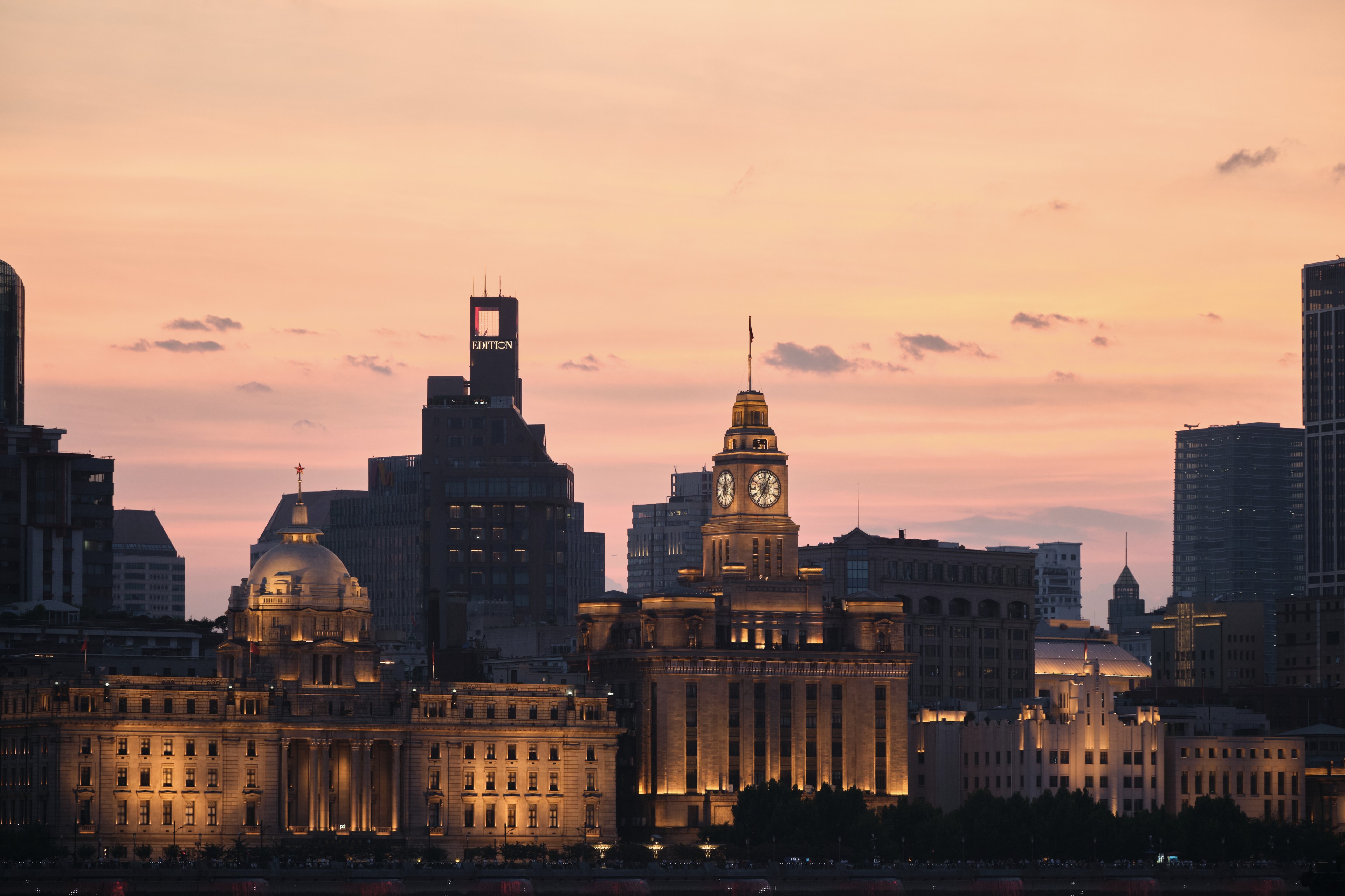 A cityscape skyline silhouettes against a pastel orange sunset with historic neoclassical buildings illuminated in the foreground
