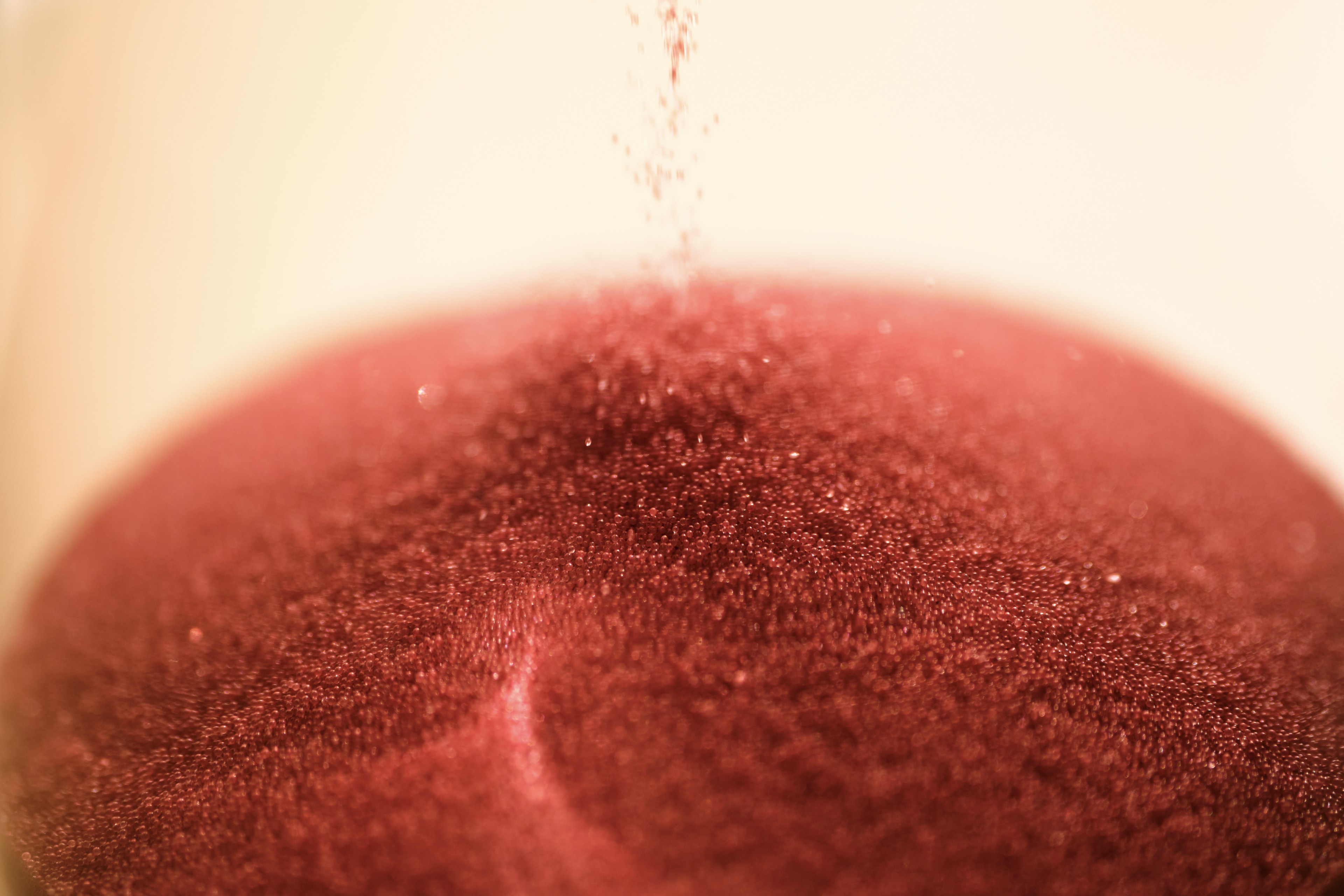 Close-up of red sand particles falling from above to form a conical mound against a blurred, light-colored background