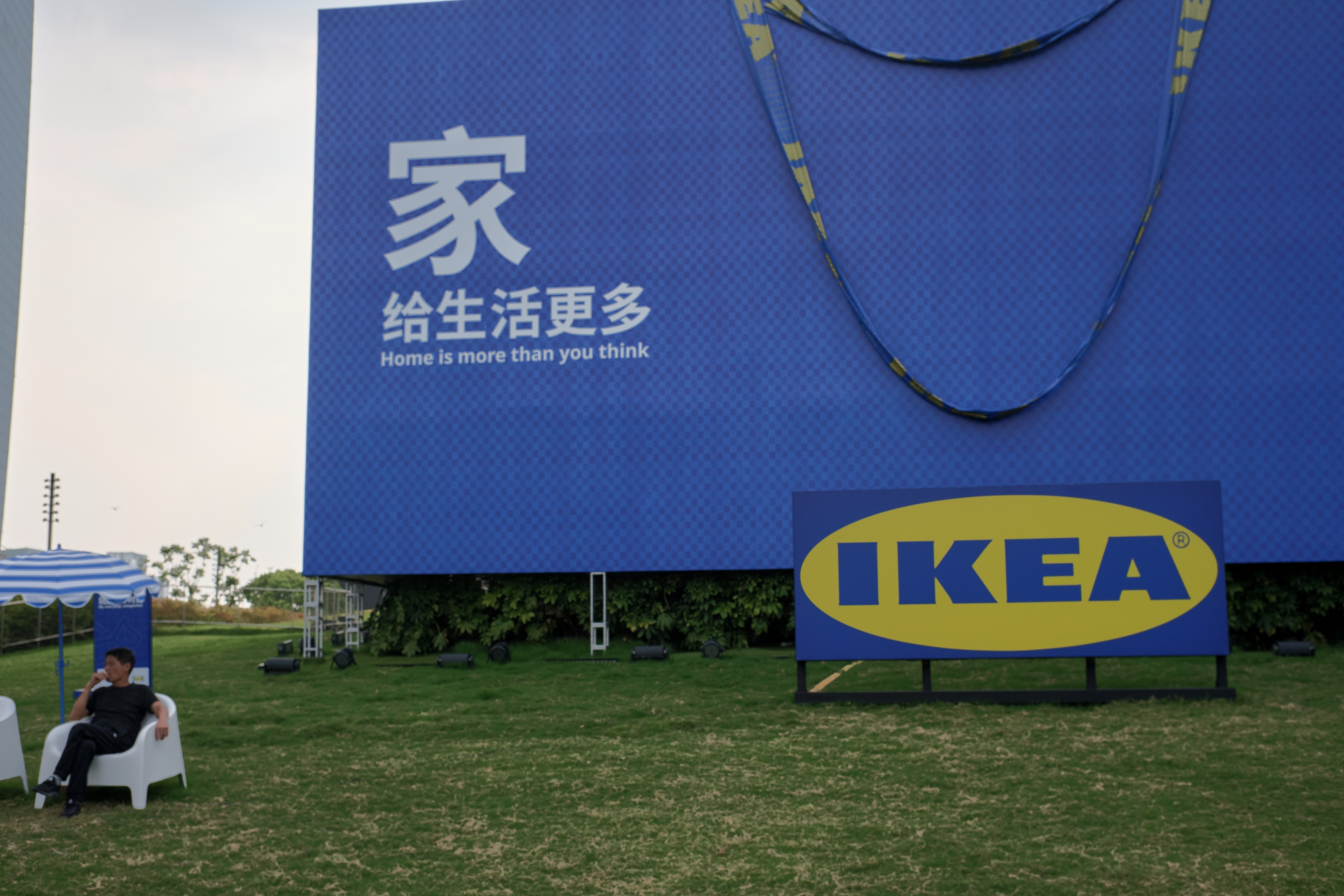 Large blue IKEA billboard with Chinese text and logo on green grass. A person sits in a white chair under a blue tent on the left