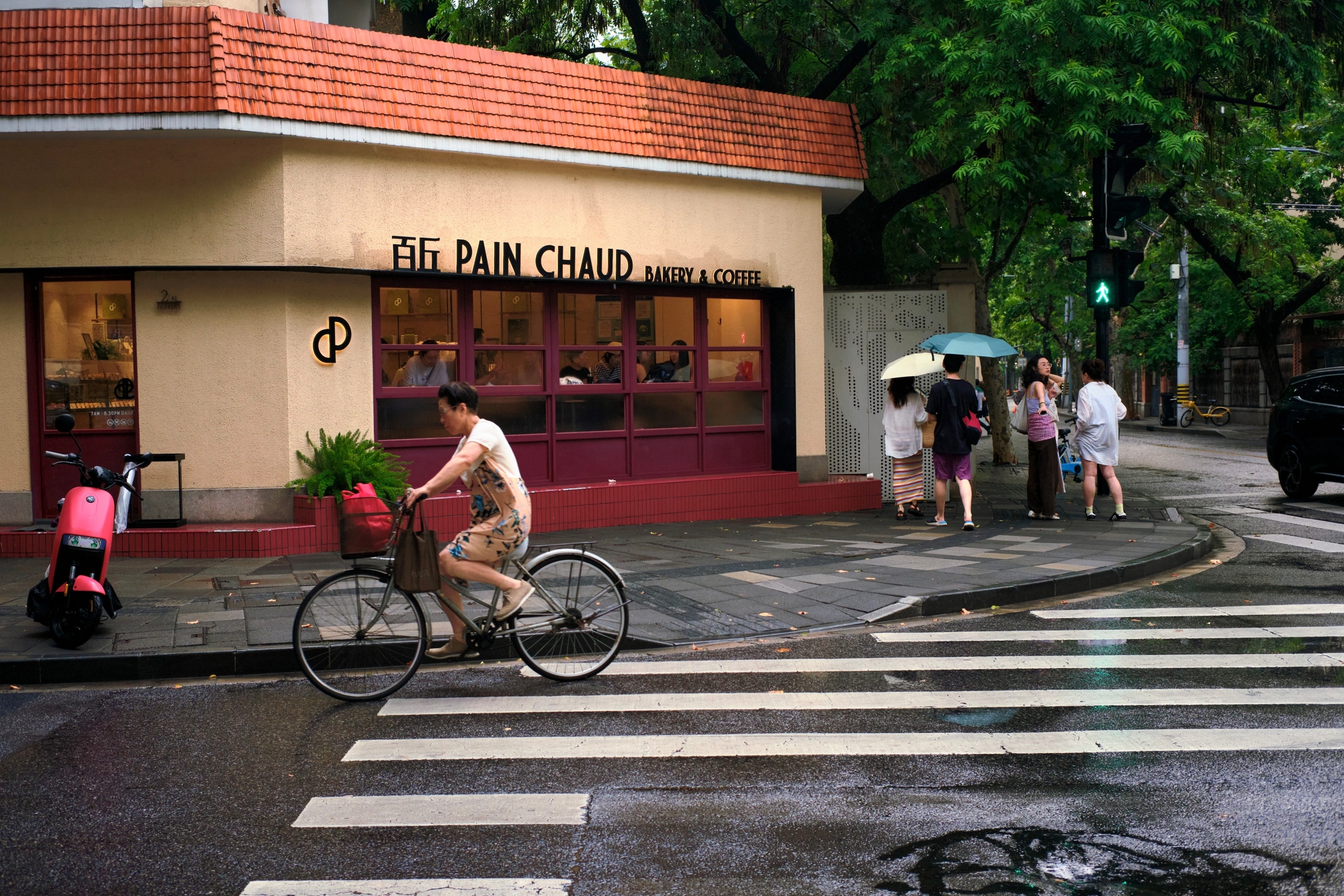 Cyclist with basket of greenery crosses a wet street on a crosswalk in front of a Pain Chaud bakery, with pedestrians holding umbrellas on the sidewalk