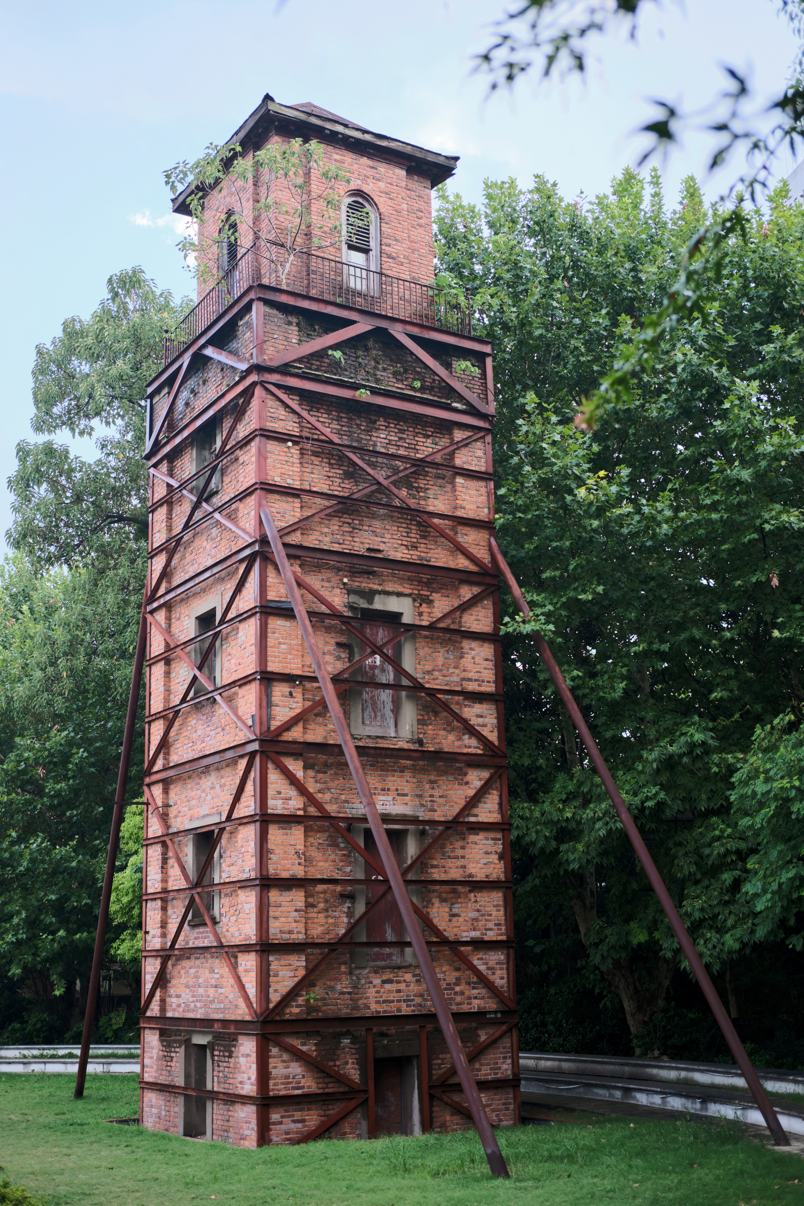 Tall brick tower with external metal framework, surrounded by green trees and grass