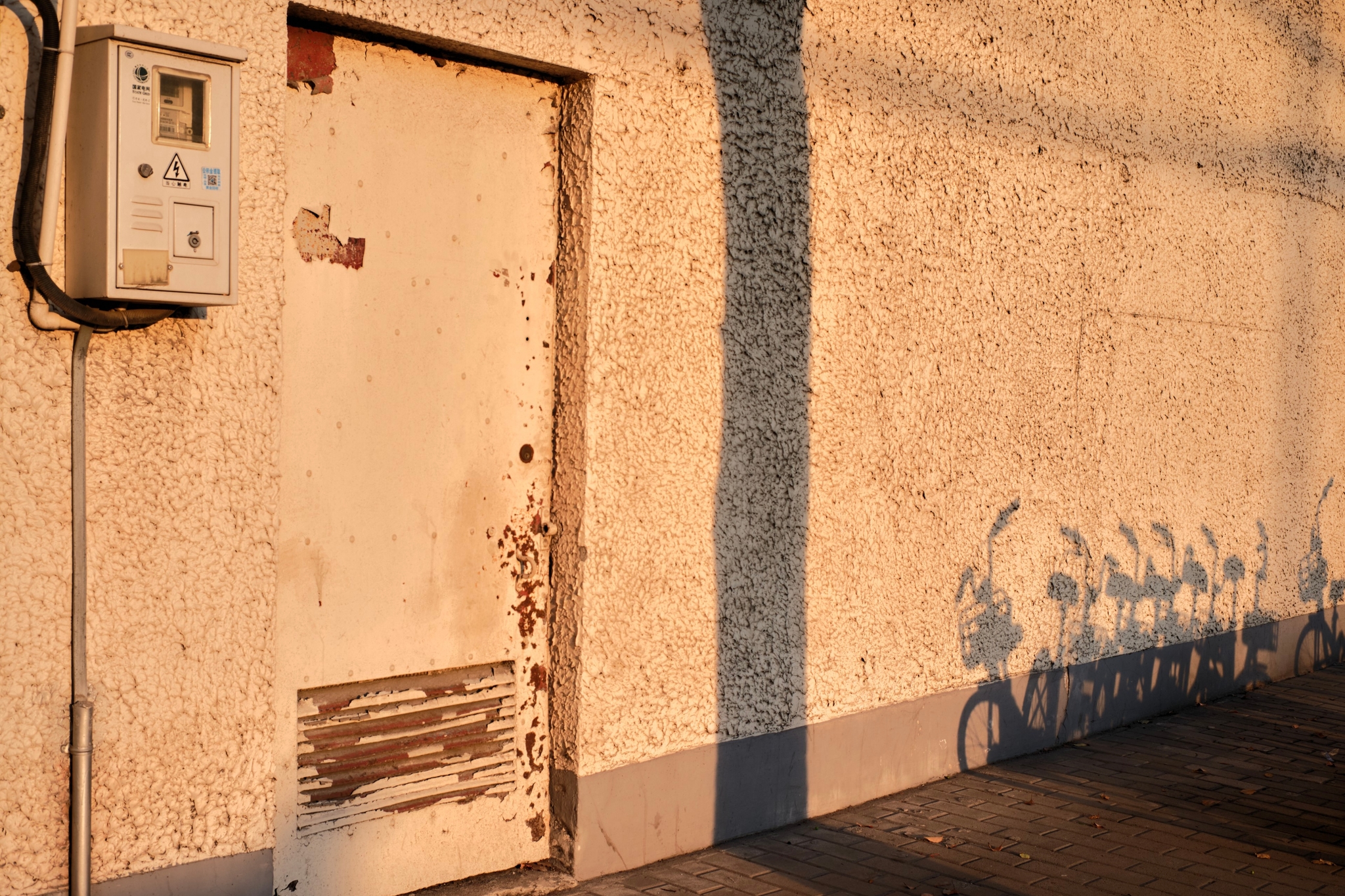 Distressed off-white door on textured light-colored wall with a utility box. Long, distinct shadows stretch across the wall and ground under warm, low-angle sunlight