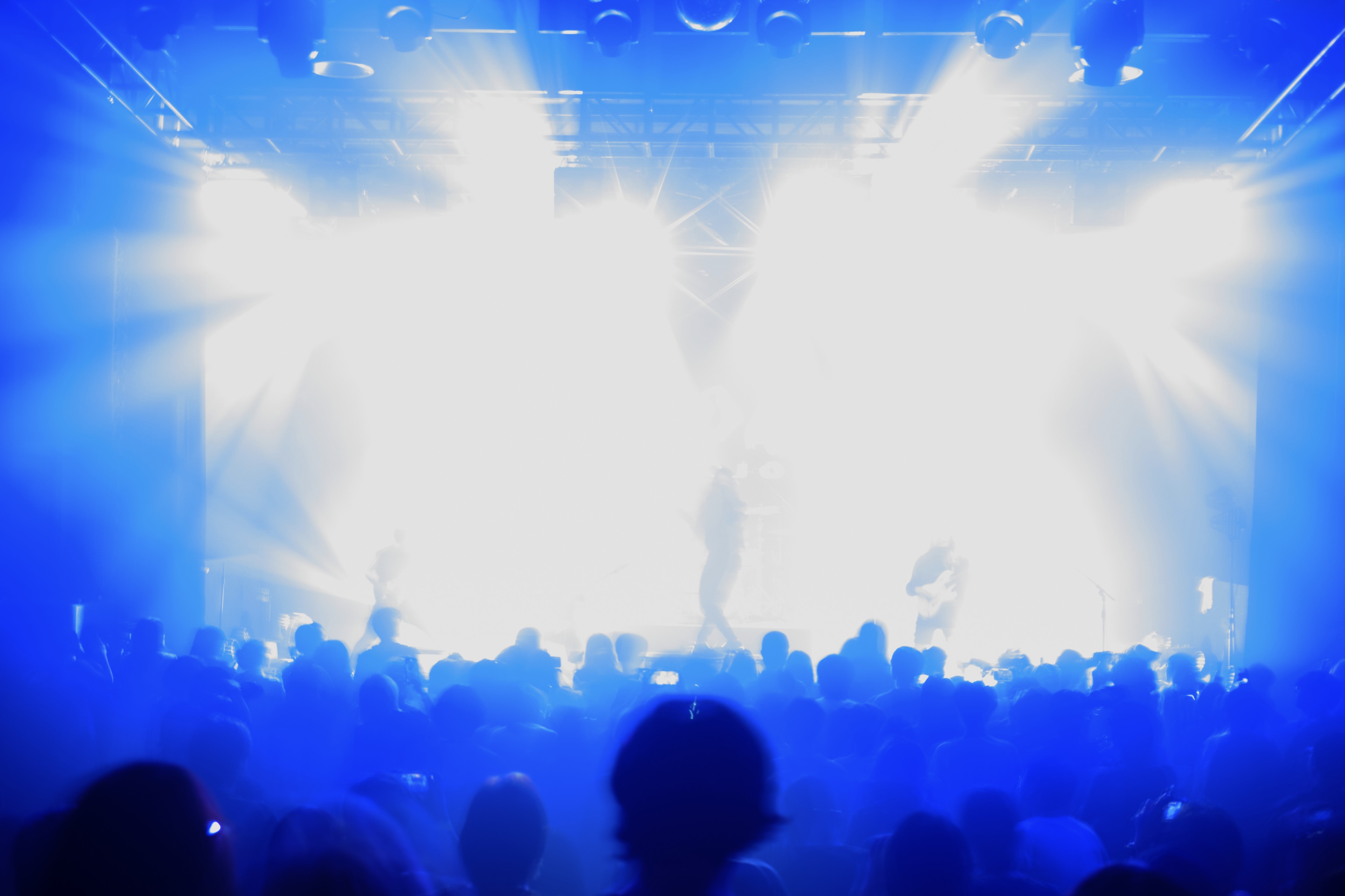 Silhouetted audience faces brightly lit stage with performers amidst blue and white spotlights