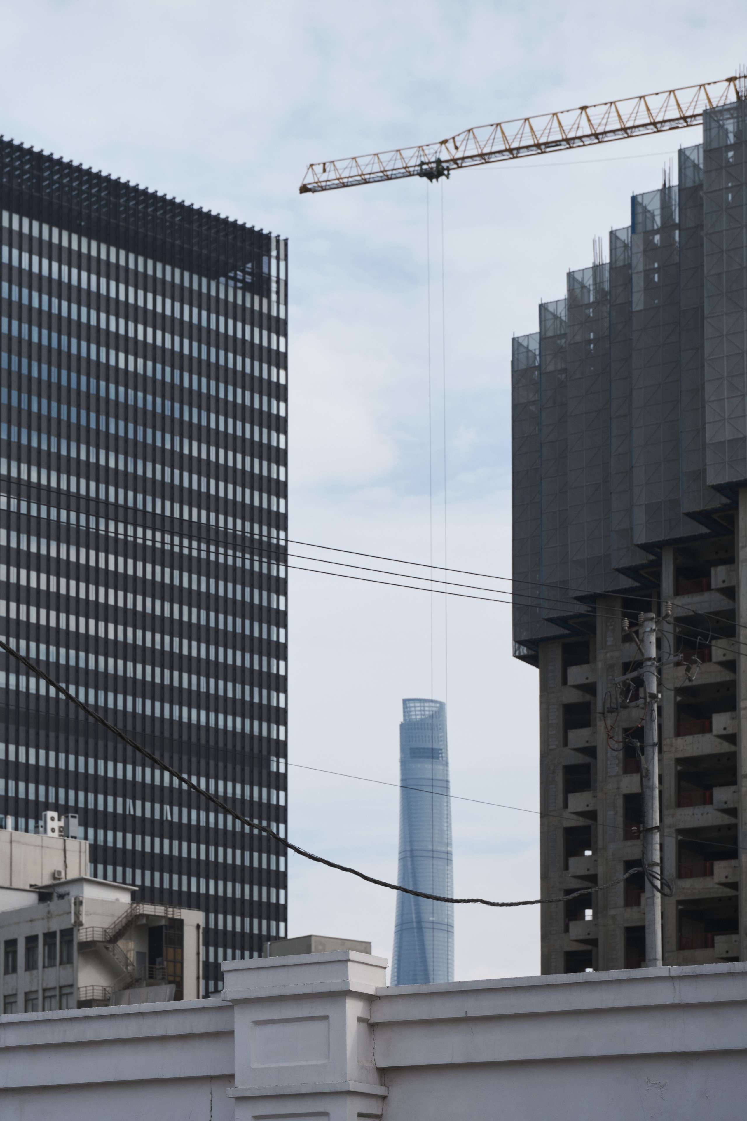 A vertical view of urban development featuring a tall office building, an unfinished skyscraper under construction with a crane, and a distant tower against a pale sky