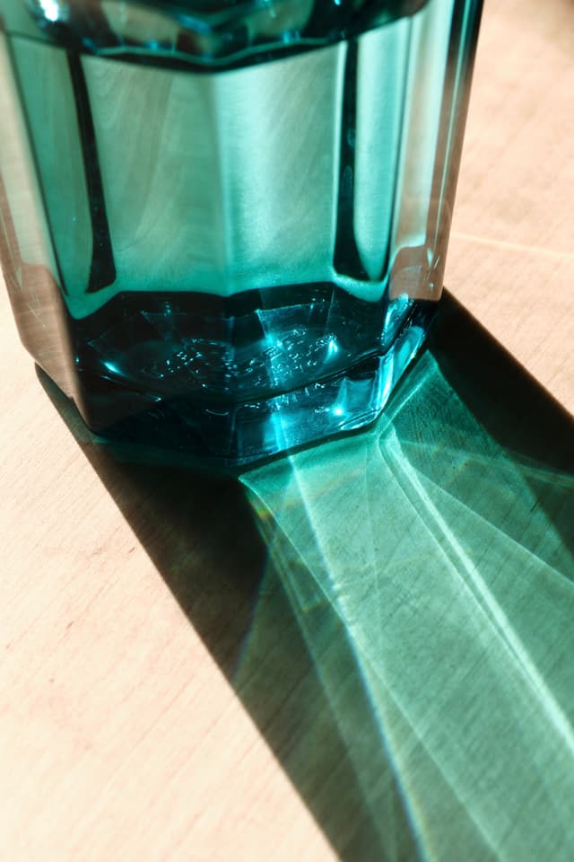 A faceted teal glass vessel sits on a light surface, casting a long, geometric shadow with distinct light refraction patterns