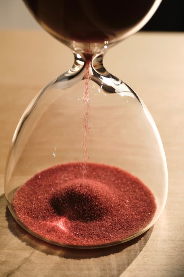 Close-up view of a glass hourglass with red sand grains falling through the narrow neck onto a mound forming in the lower bulb