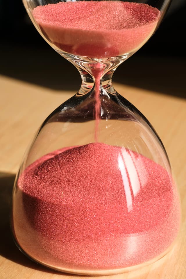 A glass hourglass filled with pink sand sits on a wooden surface, with sand flowing from the upper chamber to the lower chamber