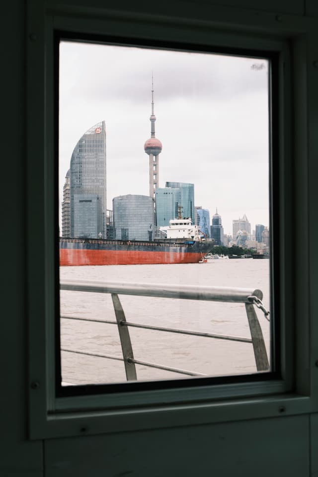 A rectangular window frame looking out over a river towards the Shanghai skyline, featuring the Oriental Pearl Tower and a large orange cargo ship passing in front of skyscrapers under a cloudy sky