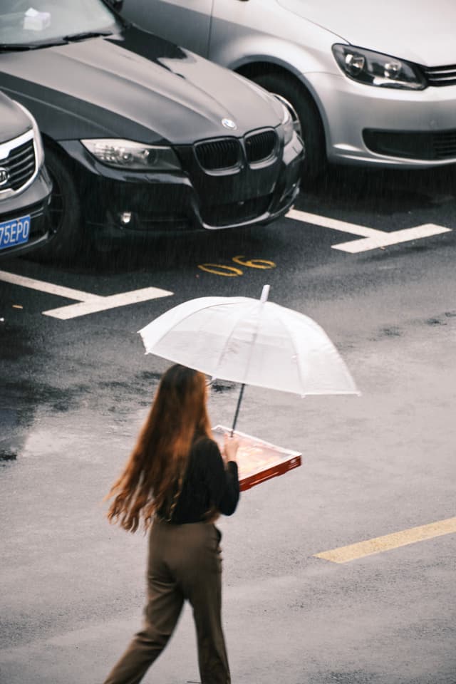 A woman with long brown hair carries a pizza box and a clear umbrella while walking across a wet, marked parking lot in front of two parked cars
