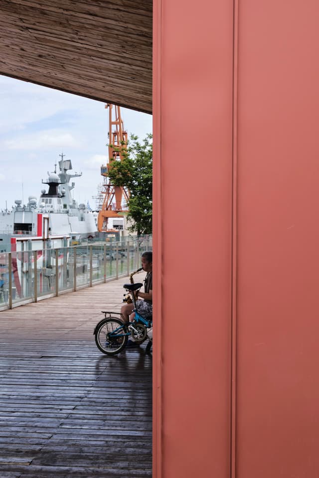 A person on a bicycle sits on a wooden pier near a railing with a large ship and orange industrial crane in the background, partially obscured by a red wall in the foreground