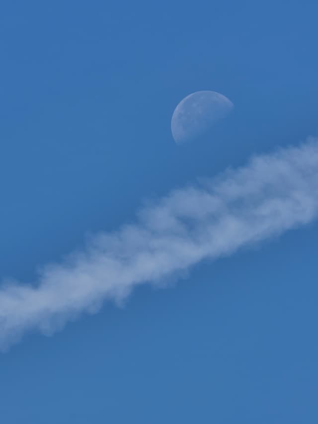 A waxing gibbous moon positioned in a clear blue sky above a diagonal contrail of white cloud vapor