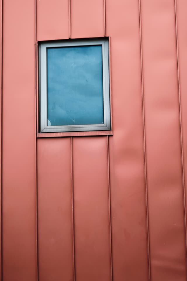 A rectangular window set into a vertical panelled wall with a dusty rose metallic finish, reflecting a cloudy blue sky