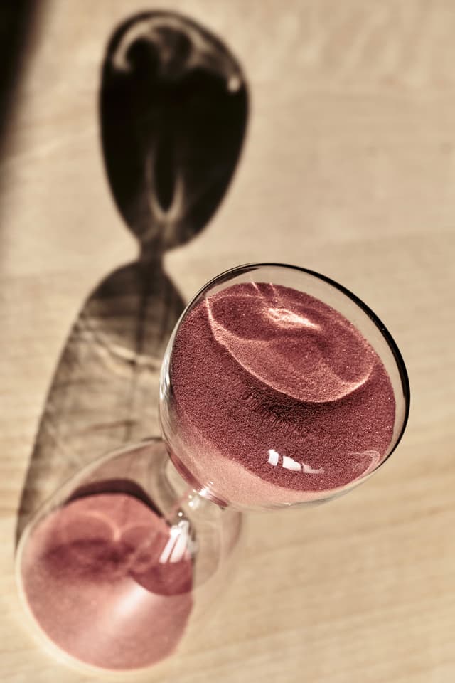 A high-angle close-up of a glass hourglass lying on its side, containing fine pink sand, casting a prominent, elongated shadow on a textured surface