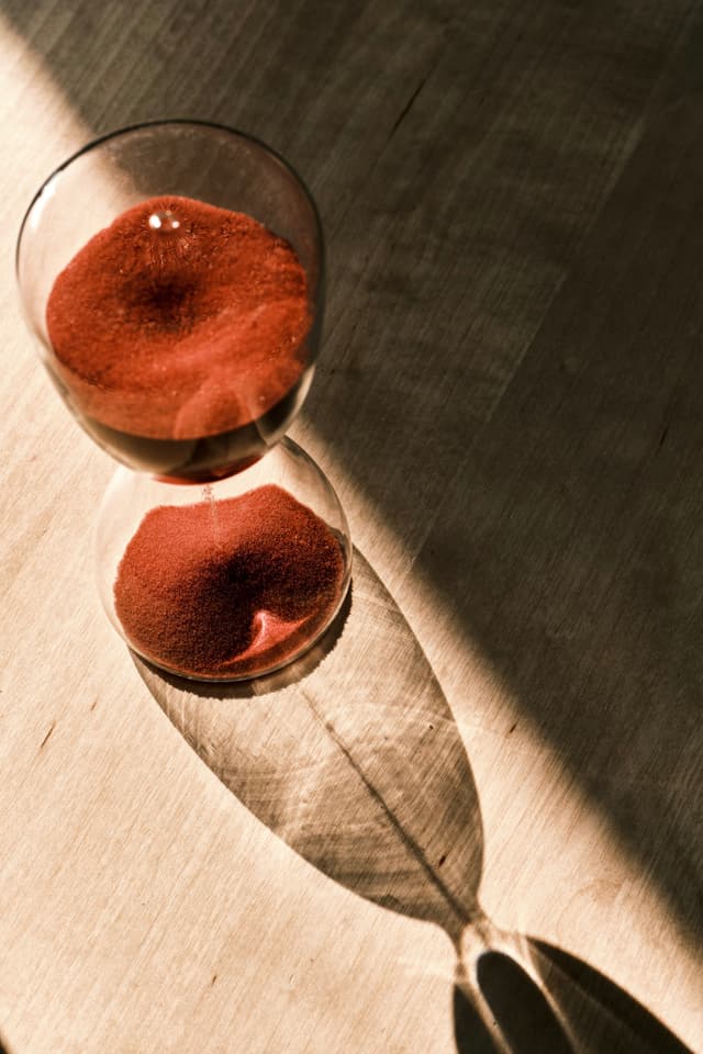 An hourglass filled with red sand sits on a wooden surface, casting a long, elongated shadow under dramatic directional lighting