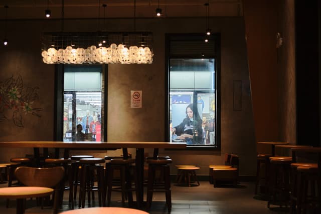 Dimly lit cafe interior with a long communal table, two windows revealing an urban scene, and a decorative chandelier