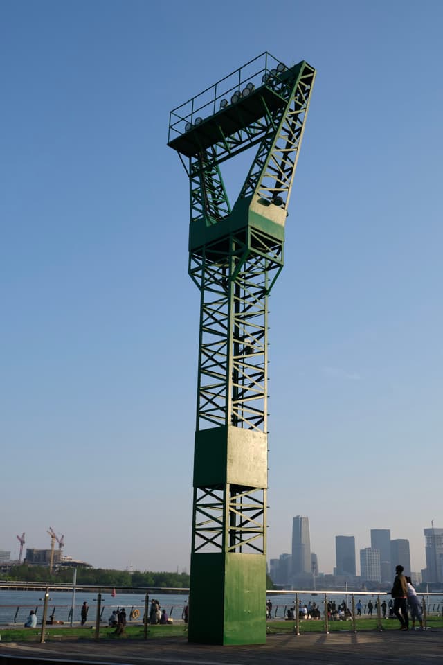 Green metal lattice tower, clear blue sky, waterfront with distant cityscape and people