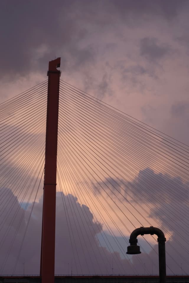 Red cable-stayed bridge pylon with radiating cables against a cloudy, multi-hued sky. Dark L-shaped pipe structure in foreground