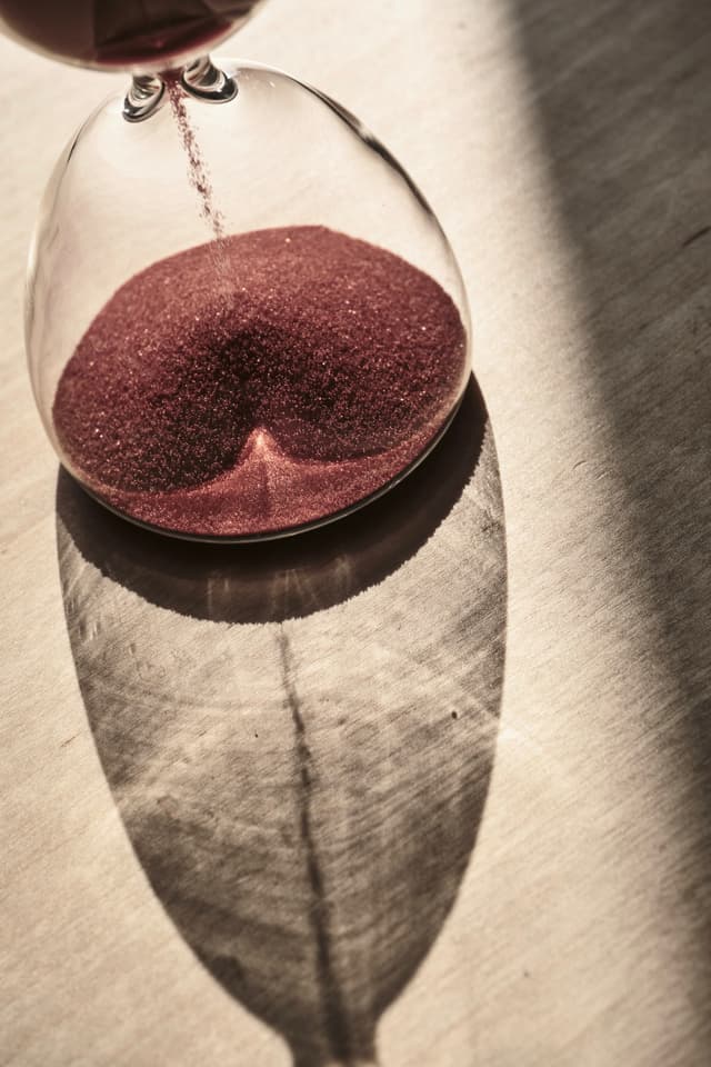 A close-up view of a glass hourglass filled with red sand sitting on a textured surface, casting a long, dark shadow in bright natural light