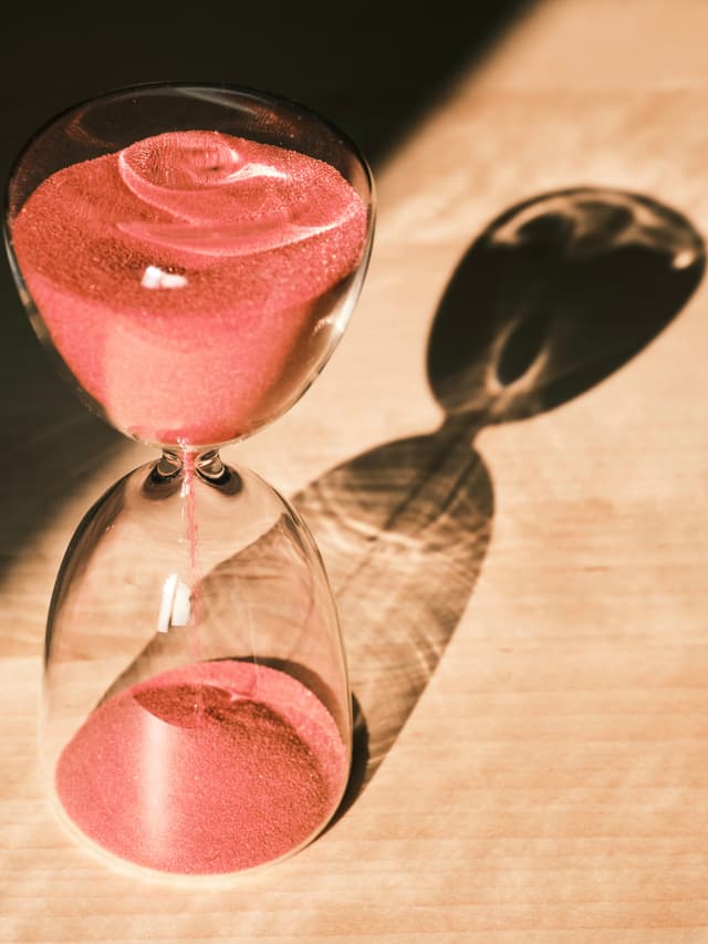 A glass hourglass containing pink sand sits on a light-colored surface under direct sunlight, casting a large elongated shadow