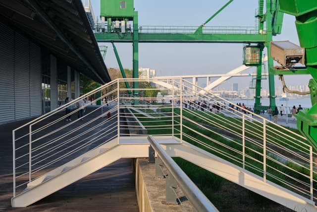 Modern metal staircase leading to waterfront, green industrial cranes, white arched bridge, building on left