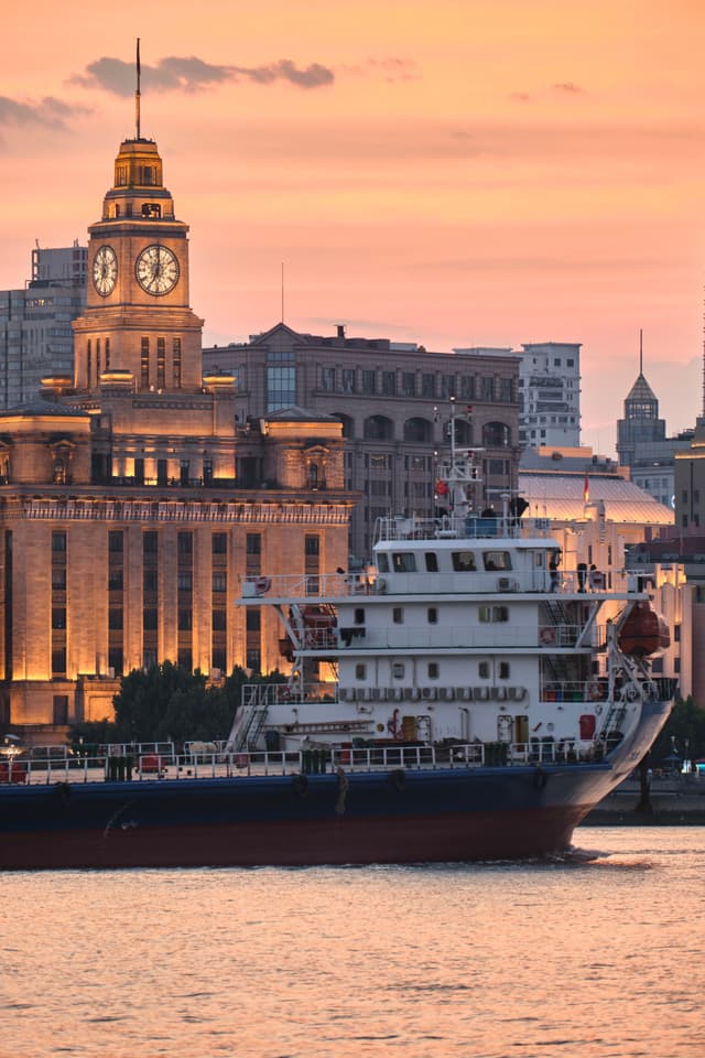 A cargo ship cruises along the Huangpu River in Shanghai past the historic clock tower of the Customs House during sunset with soft orange light reflecting on the water