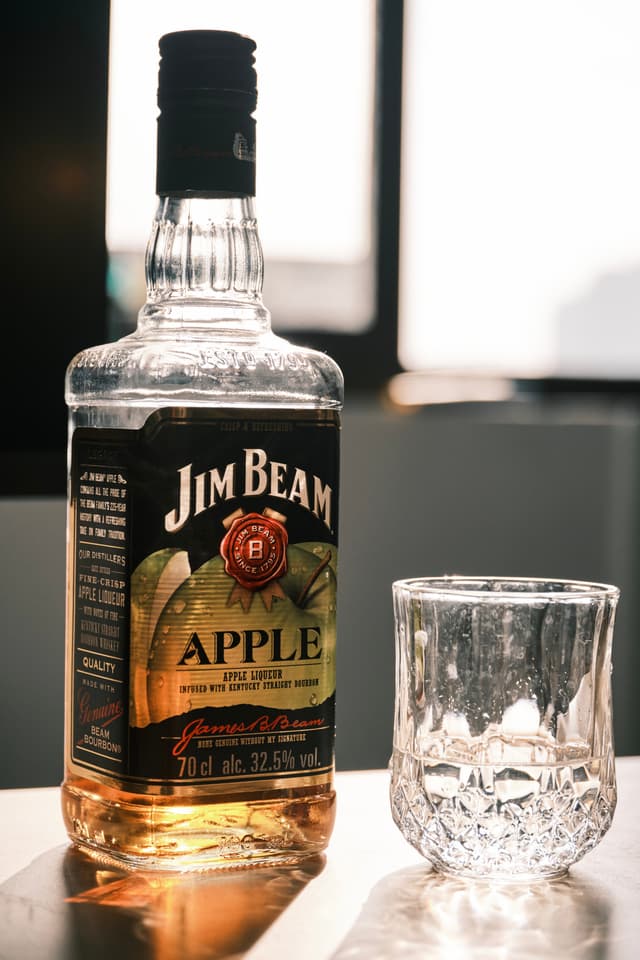 A bottle of Jim Beam Apple whiskey stands next to an empty crystal glass on a reflective surface with a window in the background